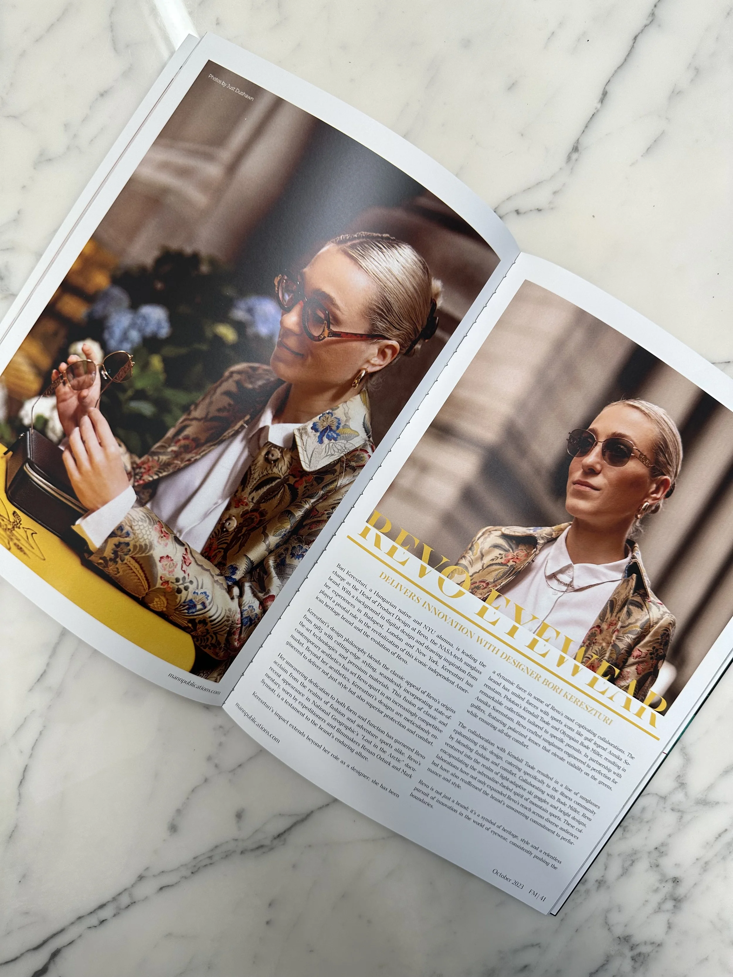 Open magazine with a fashion photograph of a woman wearing sunglasses and a floral blazer, sitting with a camera in her hand, on a white marble surface.