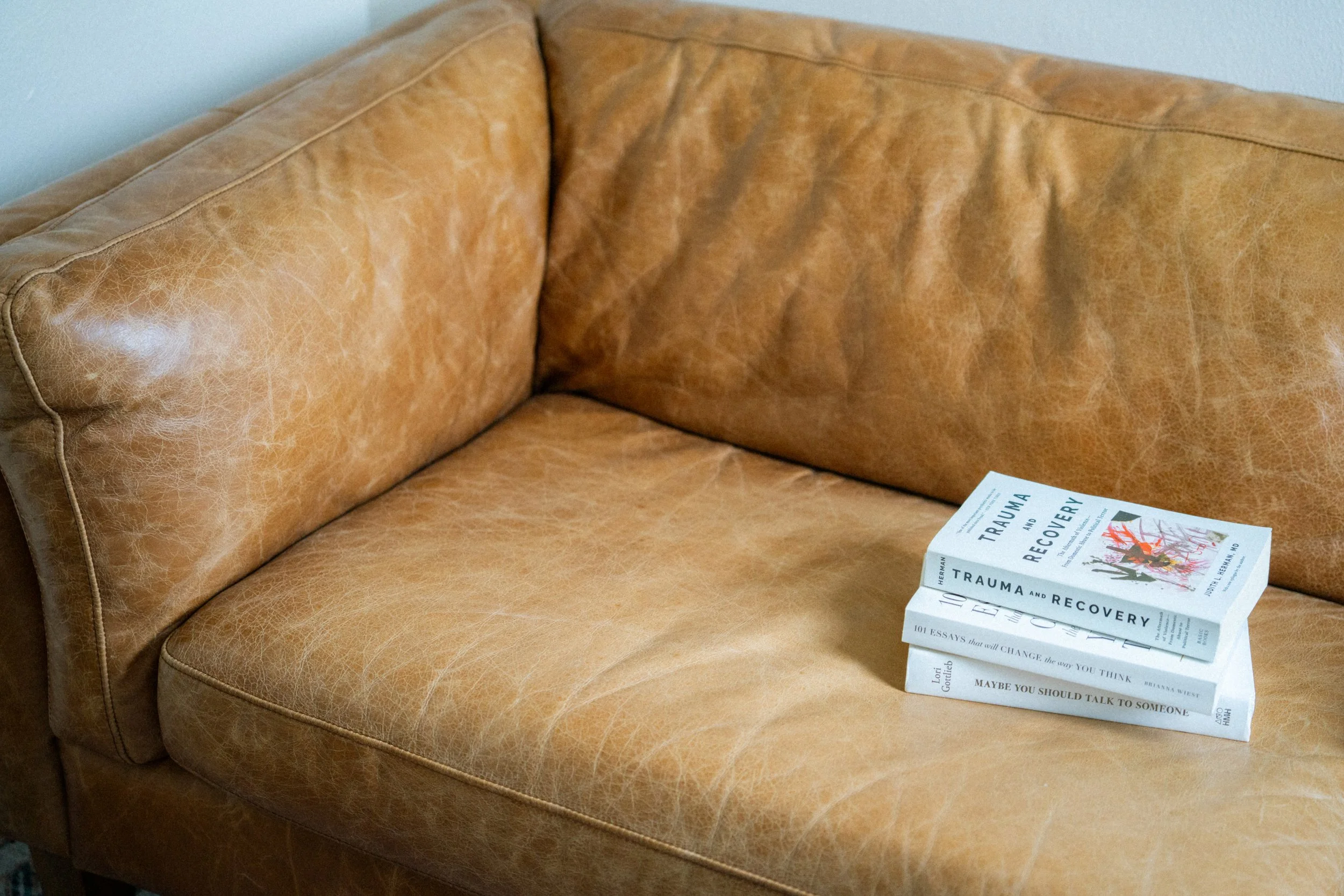 A tan leather couch with visible wear and creases, with a stack of books titled 'Trauma and Recovery' placed on the right side of the seat.