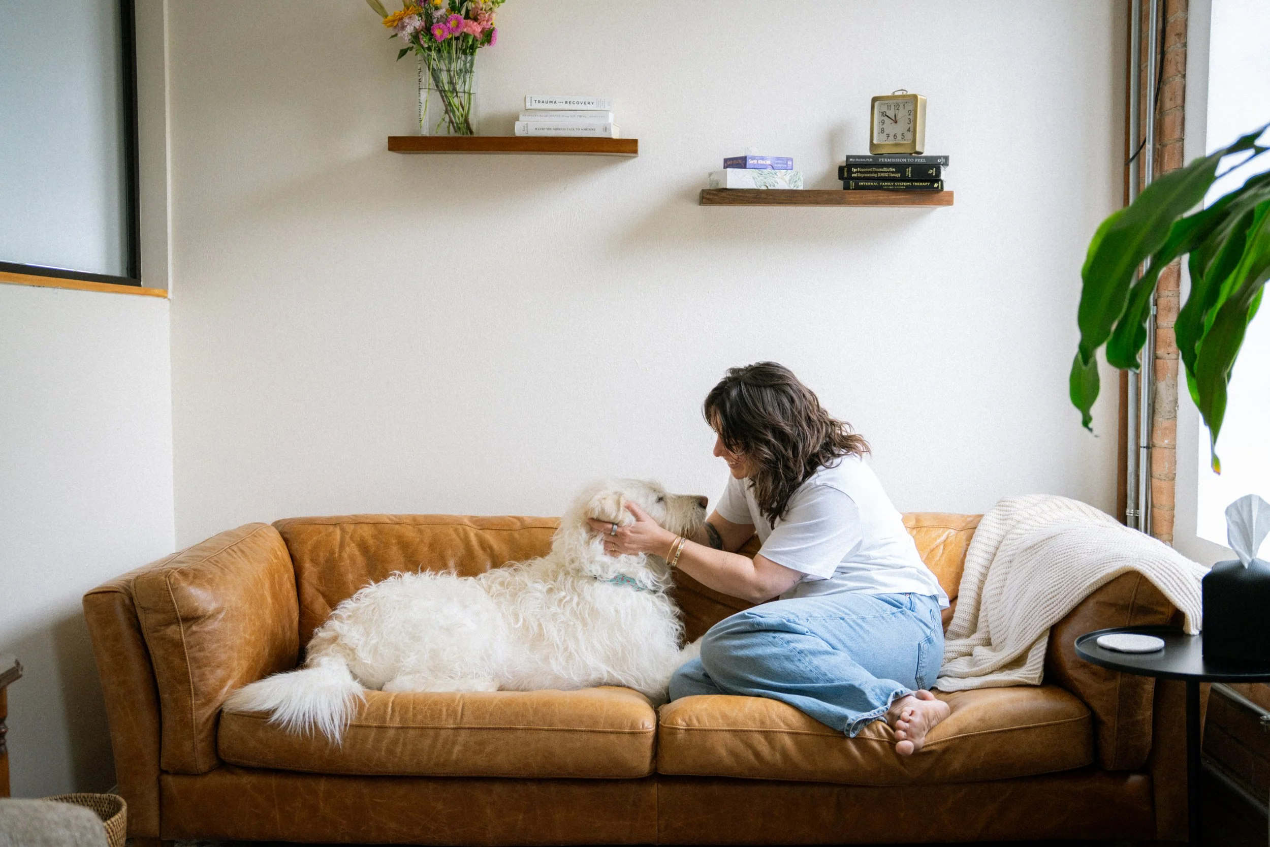 A woman sitting on a brown leather couch, petting two large white dogs in a cozy living room. The room has a white wall with two wooden shelves, a small clock, books, and a vase of colorful flowers. A large green plant is partially visible on the right.
