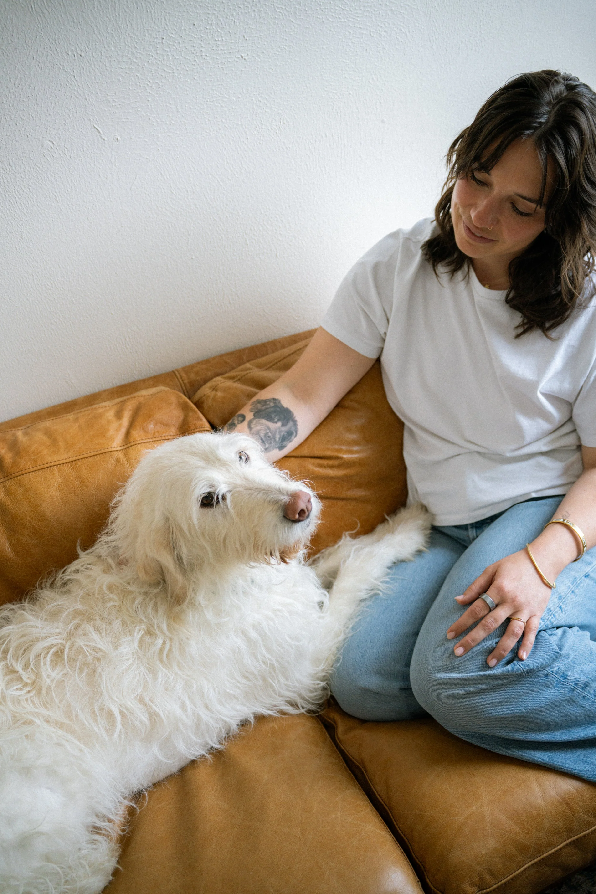 A woman with dark hair and tattoos on her arm is sitting on a brown leather couch, smiling and petting a large, fluffy white dog that is lying beside her.