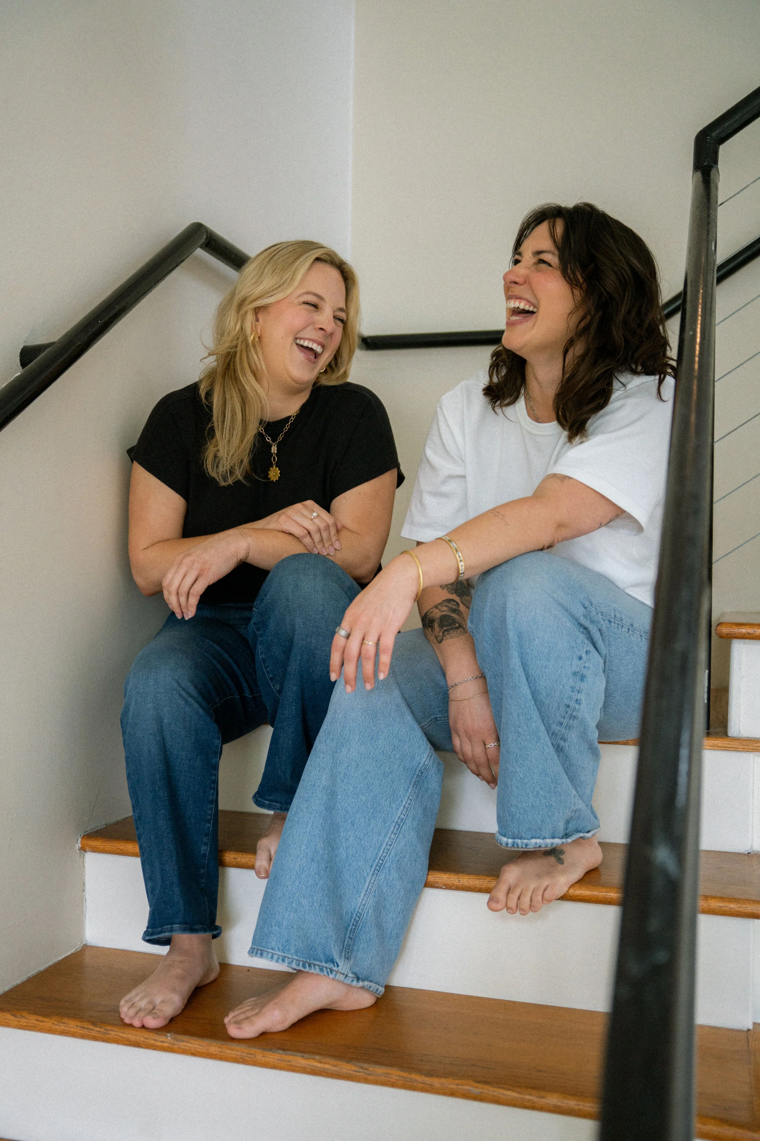 Two women are sitting on the stairs, laughing and enjoying each other's company, barefoot, with one wearing a black shirt and the other in a white shirt.