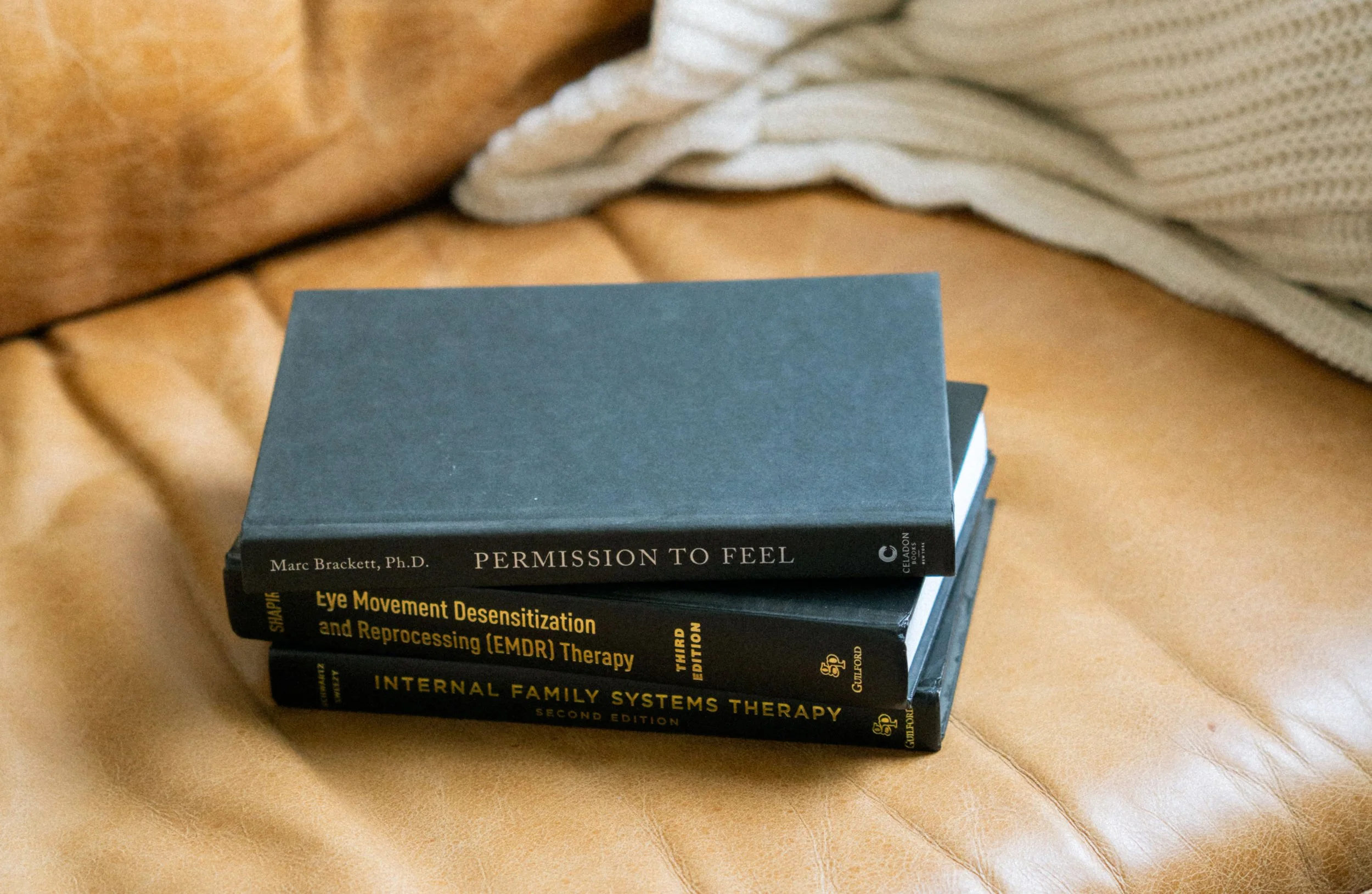 A stack of books on a beige couch, with the top book titled 'Permission to Feel' by Marc Brackett, Ph.D., and other books about eye movement desensitization and reprocessing therapy, and internal family systems therapy.