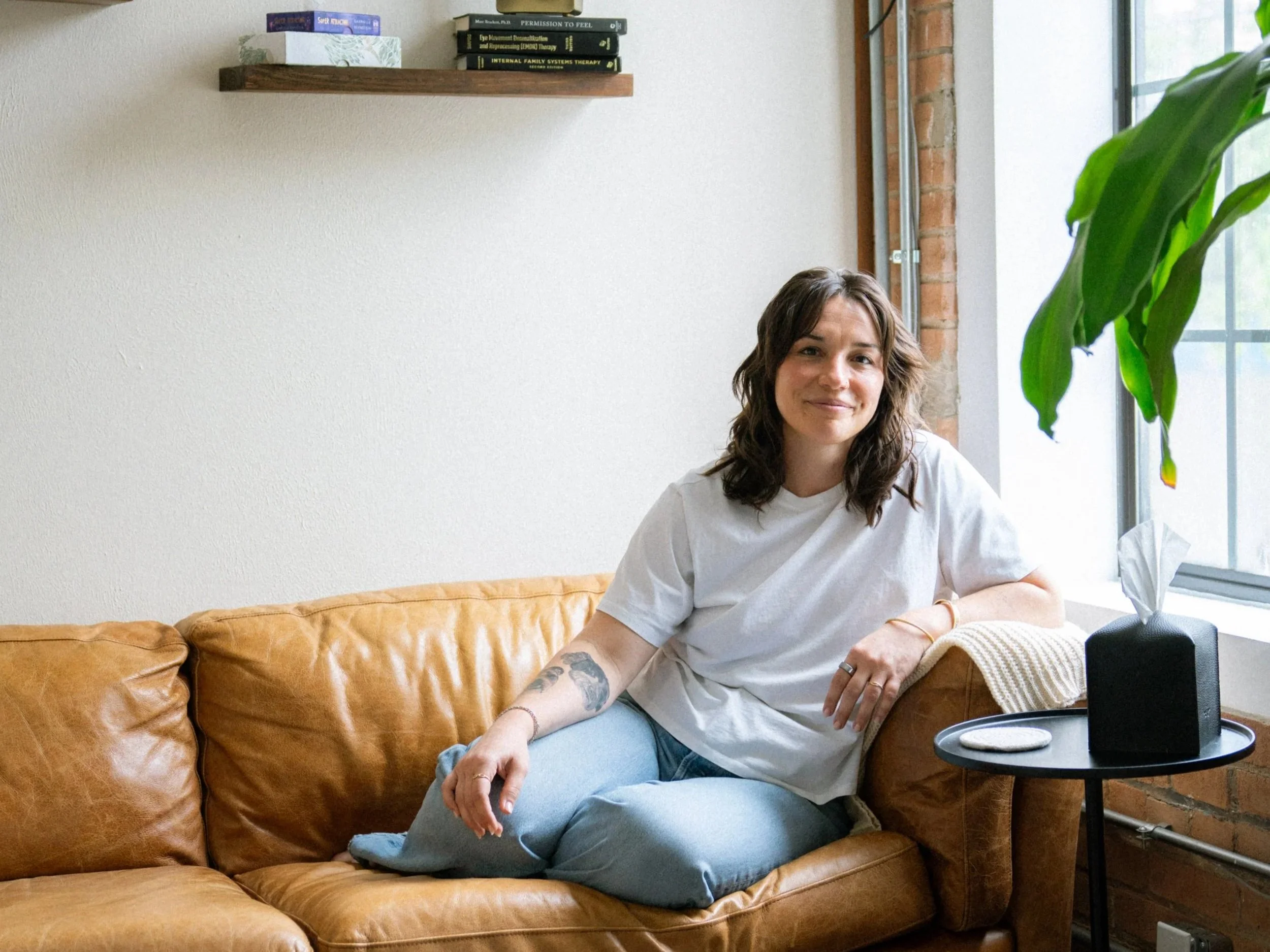 A young woman with wavy brown hair and tattoos on her arm, sitting on a tan leather couch near a window in a room with a brick wall and wooden shelf with books, smiling at the camera.