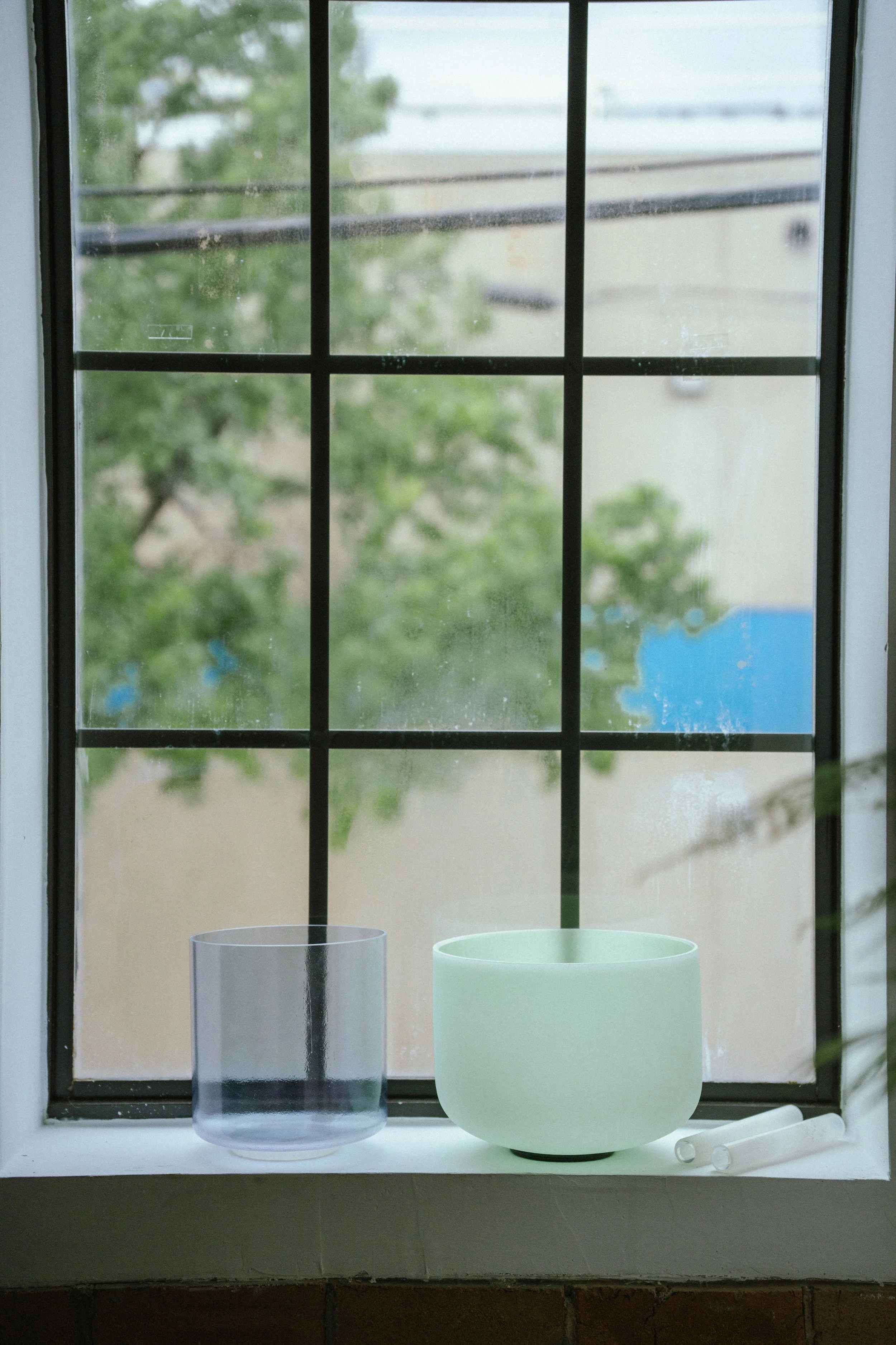 A window with a black grid, two cylindrical containers on a white windowsill, one clear and one frosted, and a white rolling pin.