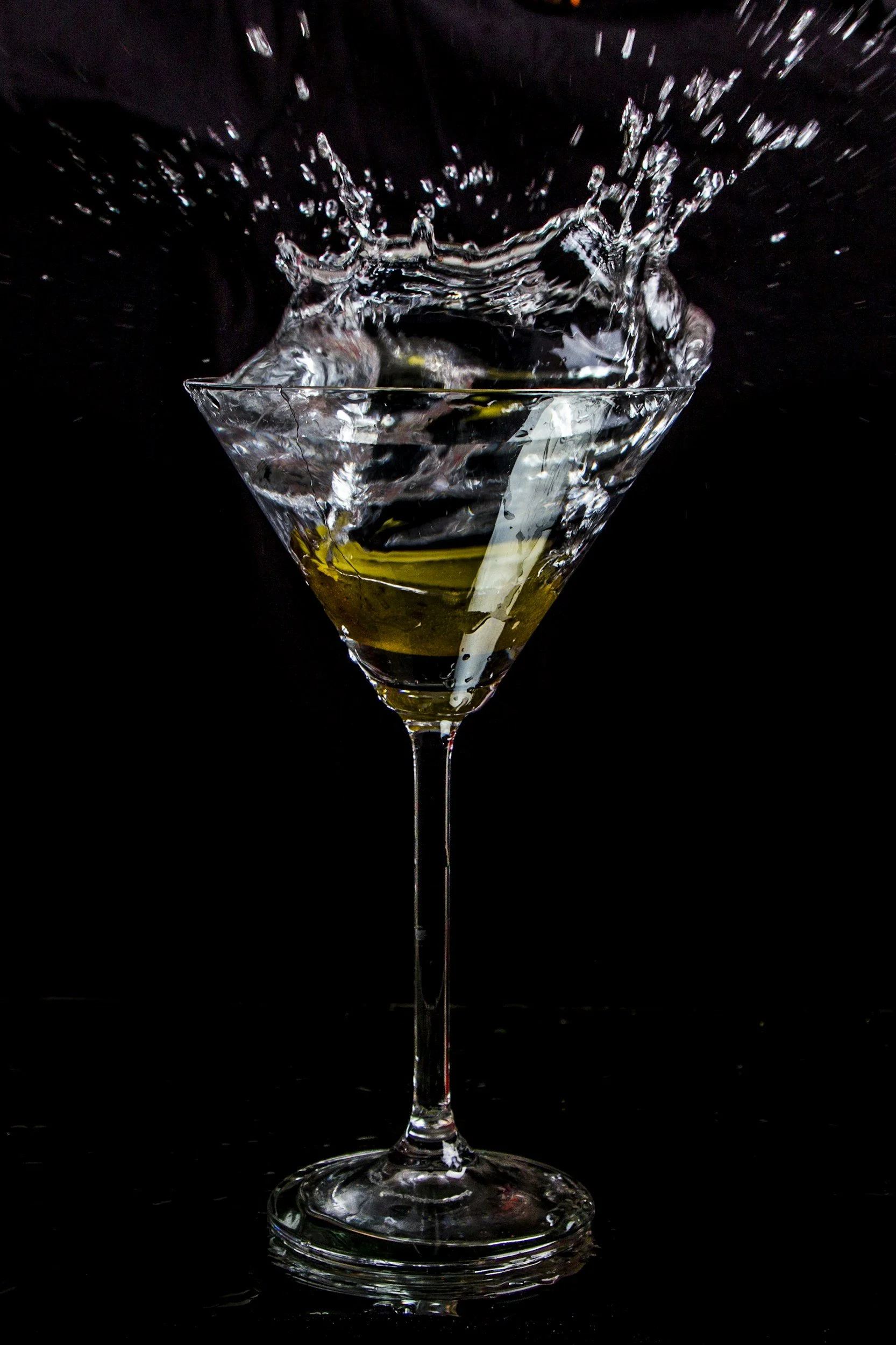 A martini glass with a splash of water and an olive, against a black background.