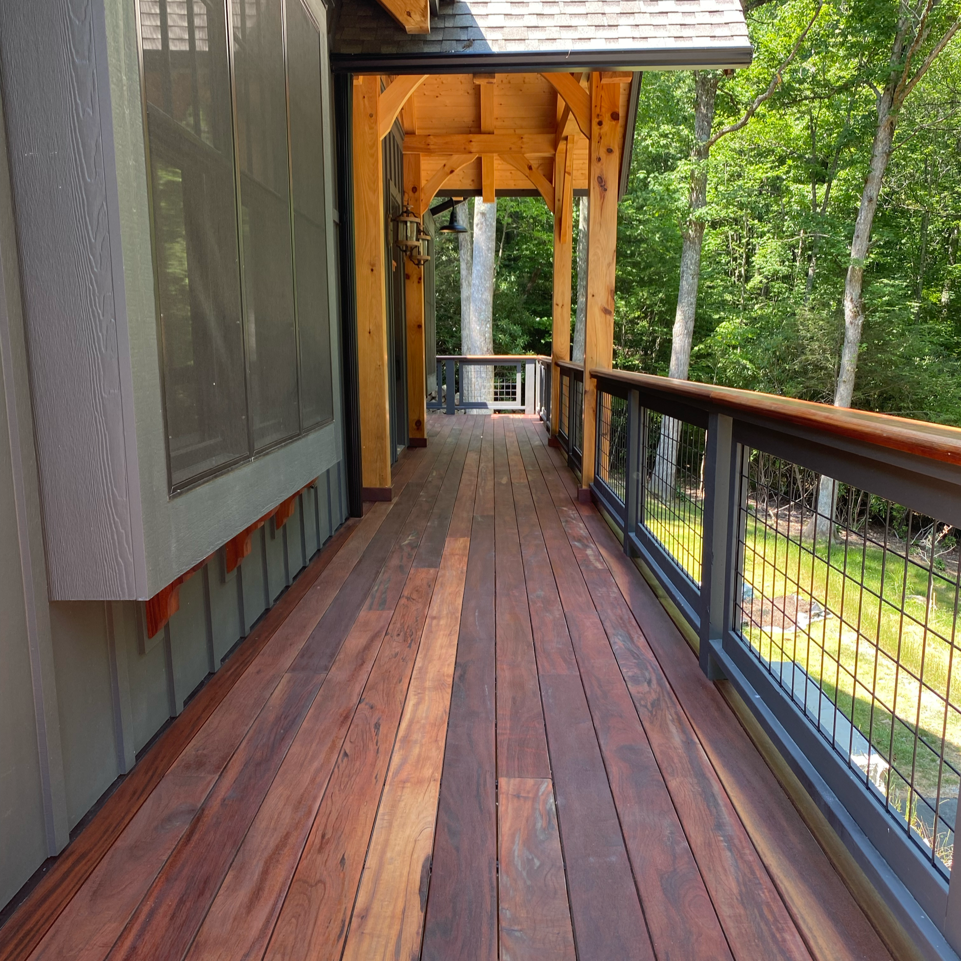 A wooden balcony with a brick-colored wooden floor, surrounded by a black and copper railing, with lush green trees in the background. The balcony has a wooden pergola overhead, part of a house with gray siding and black window frames.