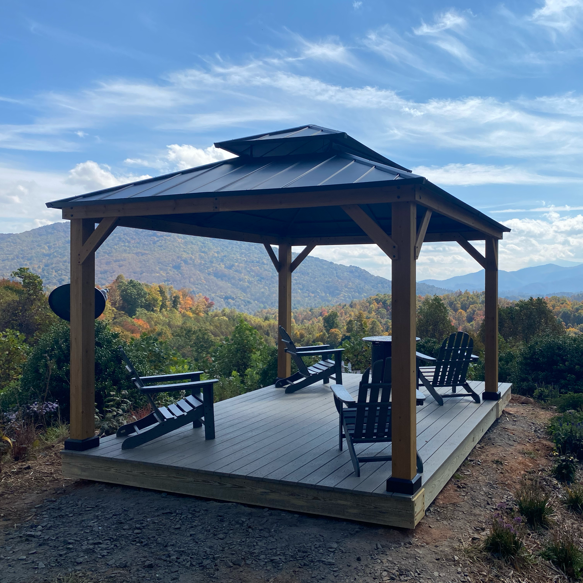 A wooden gazebo with a metal roof in a scenic mountainous area with trees and blue sky in the background, outdoor seating underneath.