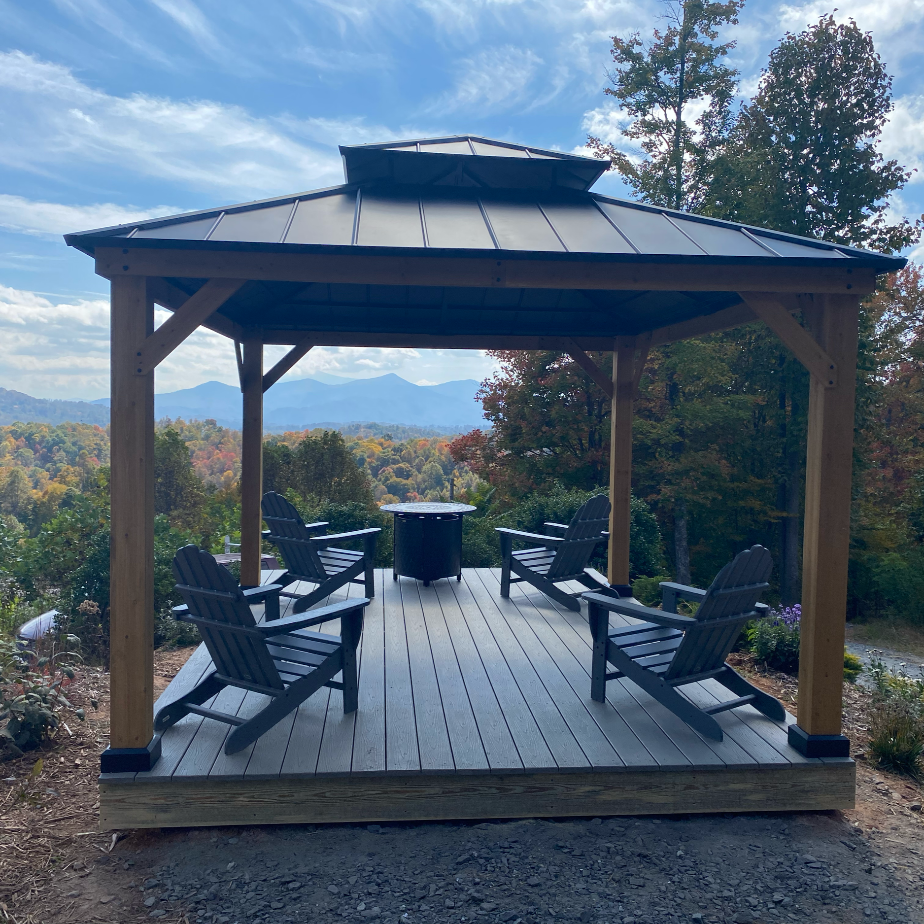 A wooden outdoor gazebo with a metal roof, surrounded by trees and mountains in the distance, with a table and chairs inside.