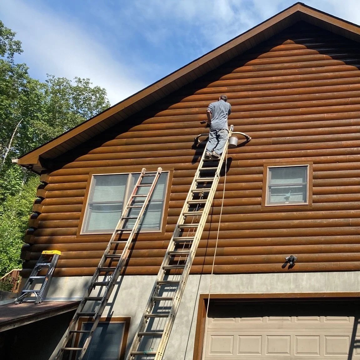 A worker standing on a tall wooden ladder cleaning the side of a two-story log cabin with a pressure washer under a partly cloudy sky.