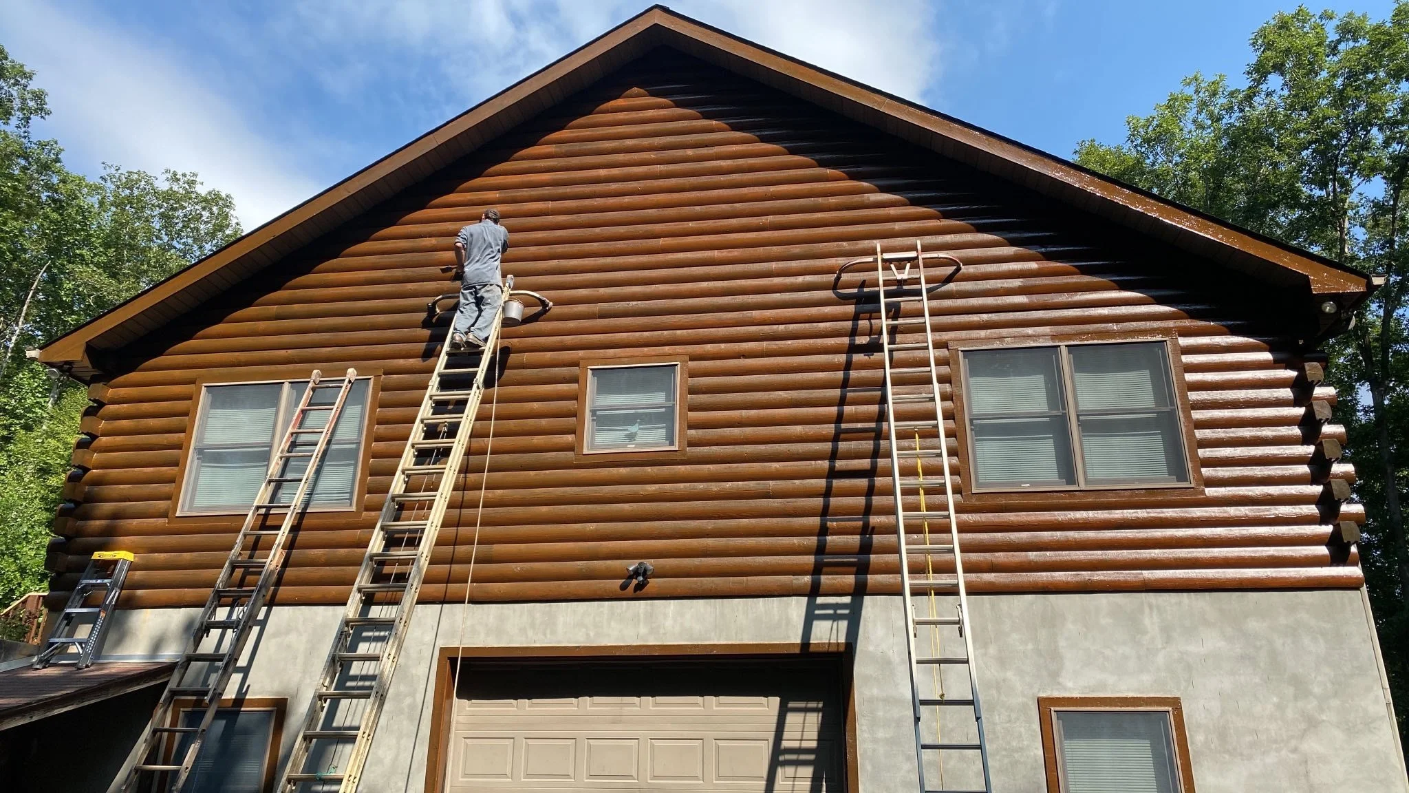A person on a ladder cleaning the exterior of a large wooden house with a Log cabin style. There are two other ladders leaning against the house, and the house has several windows and a garage door. The sky is blue with some clouds, and trees are visible in the background.