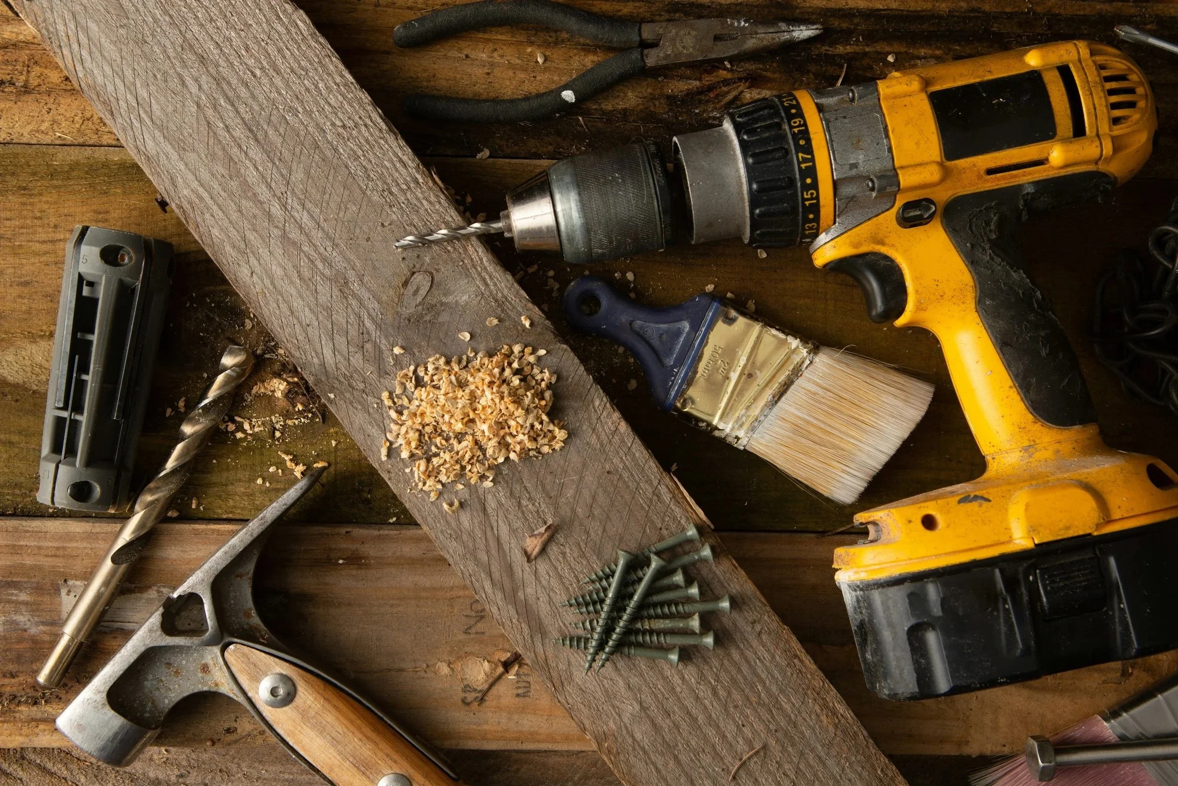 A cluttered workbench with tools including a cordless drill, drill bits, screws, a paintbrush, and a paint scraper, along with wood shavings and nails scattered on the surface.