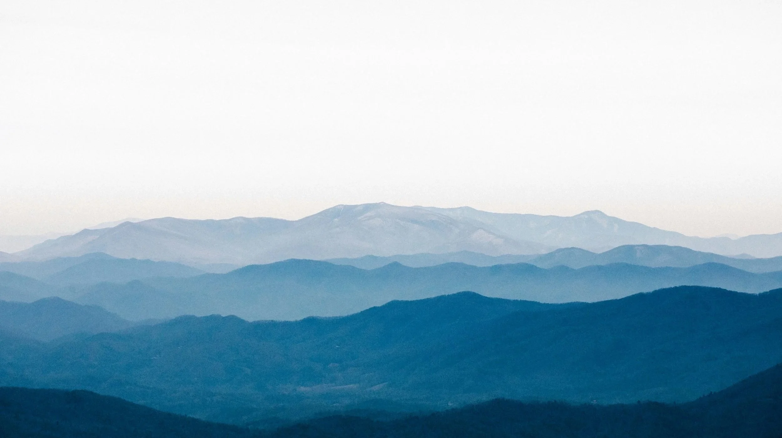 A layered mountain range under a pale, overcast sky with blue and gray tones.