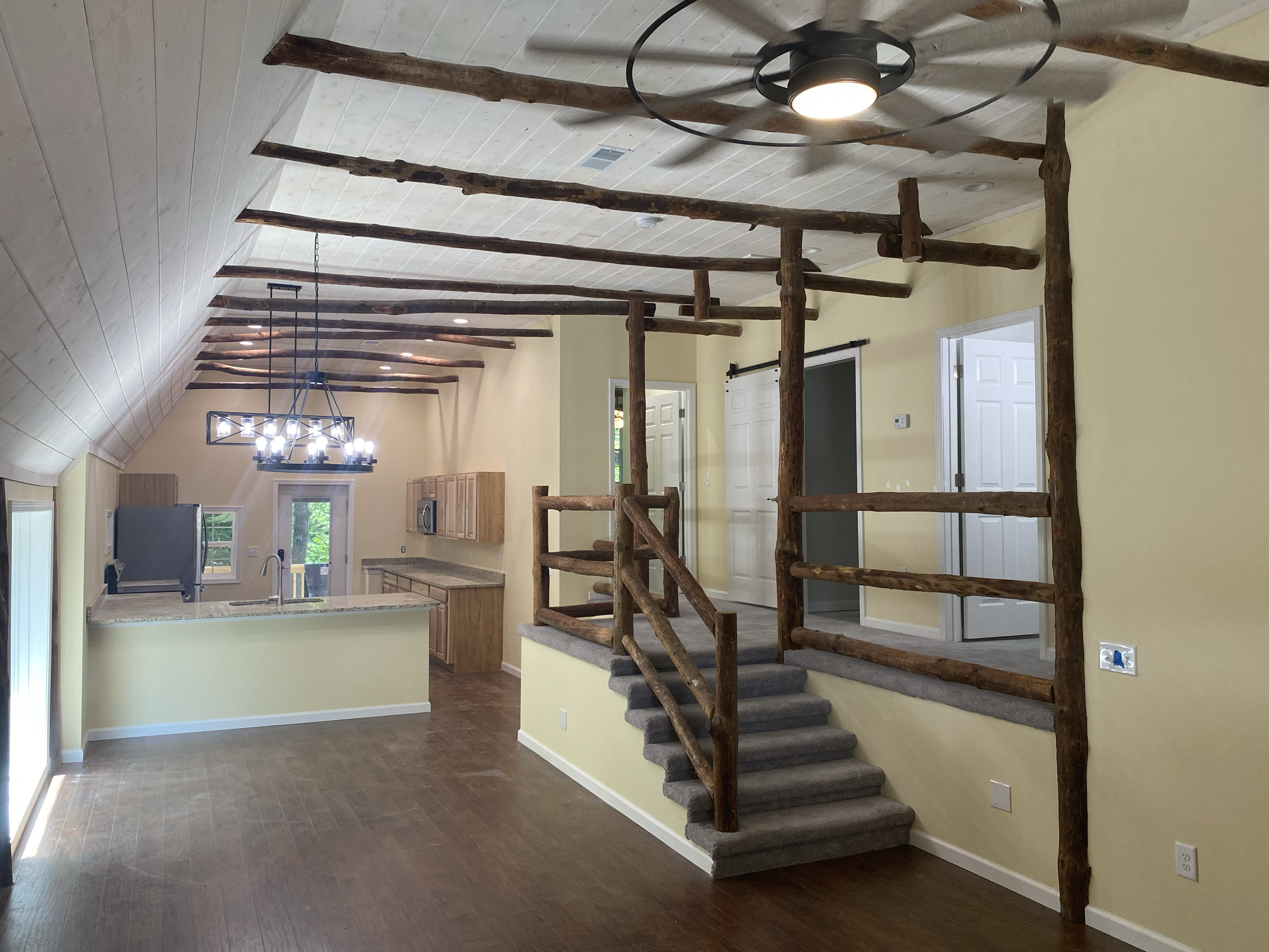 Interior of a house showing a staircase with rustic wooden handrails and steps covered with gray carpet, leading to a split-level area with light yellow walls, white doors, and a kitchen in the background. Modern ceiling fans and a chandelier are als