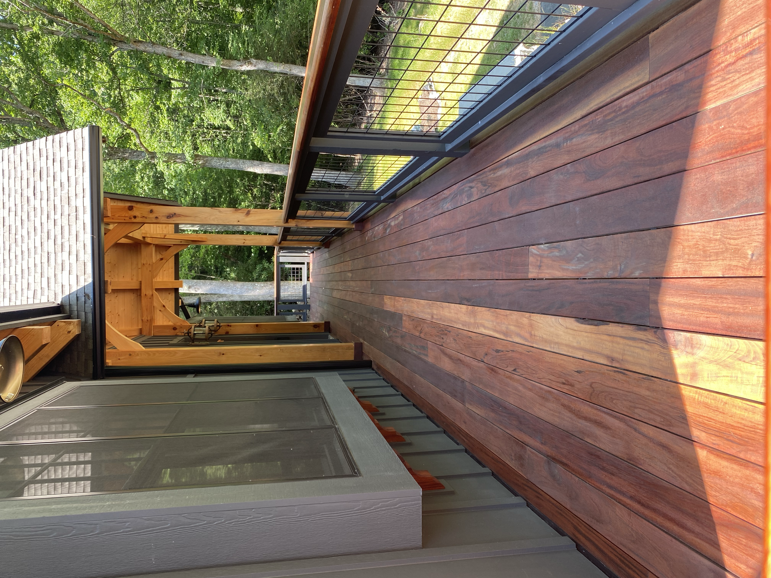 A wooden deck with reddish-brown planks adjacent to a house with gray siding, surrounded by green trees and grass, and a black metal railing.
