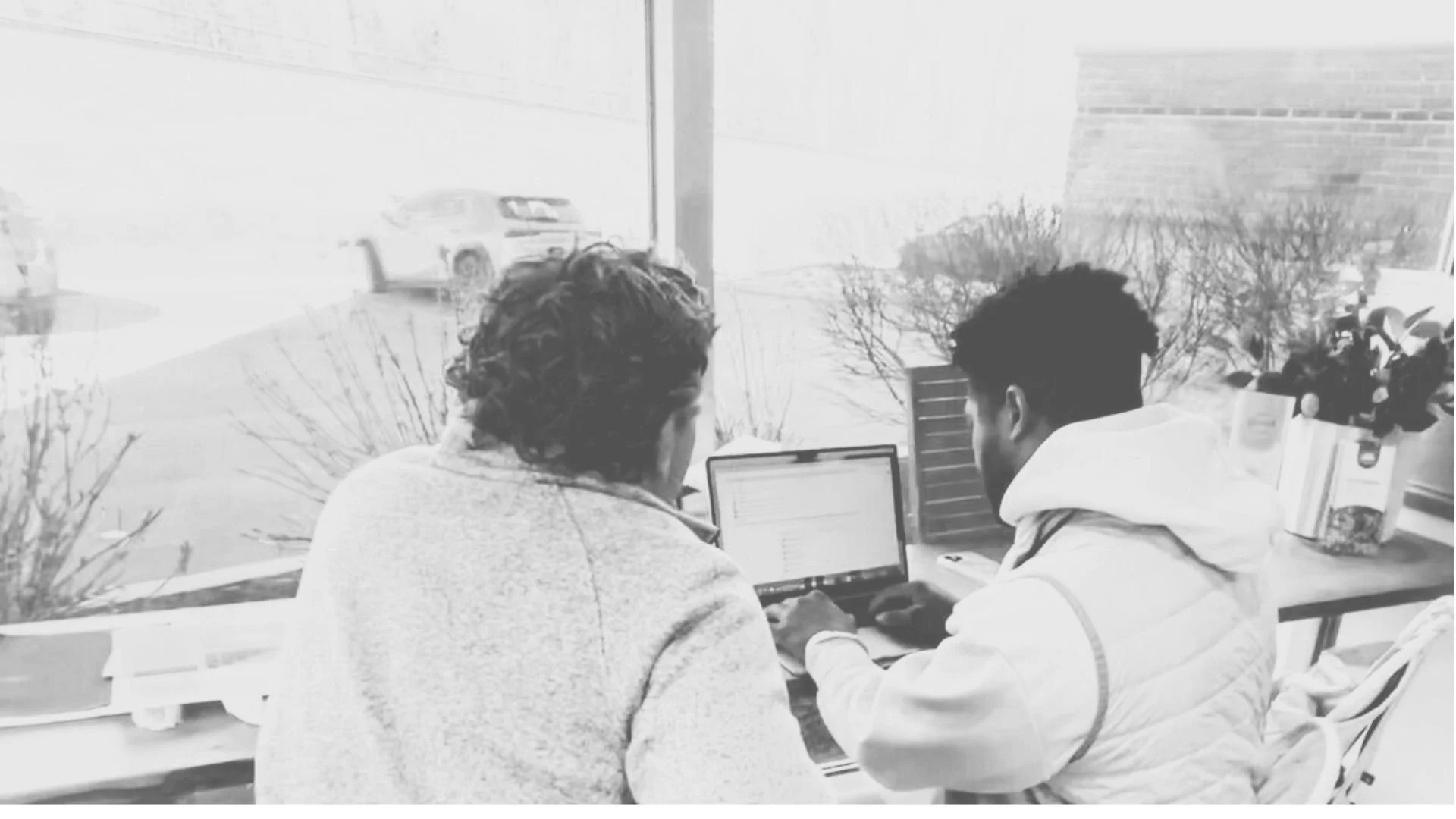 Two individuals sitting at a table in front of a large window, working on a laptop, with cars visible outside.