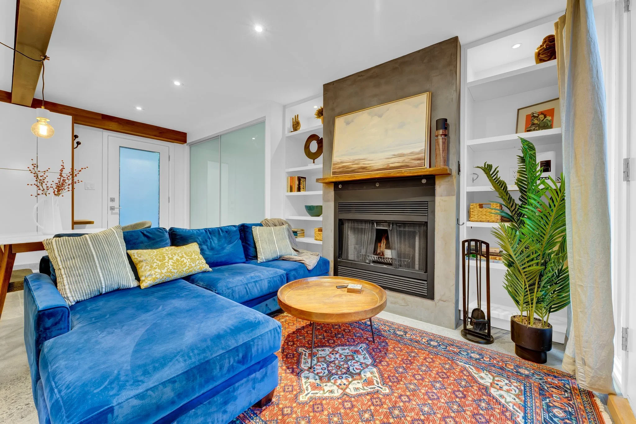 Living room with blue velvet sofa, patterned pillows, fireplace, built-in white shelves, potted plant, and a wooden coffee table on an orange patterned rug.