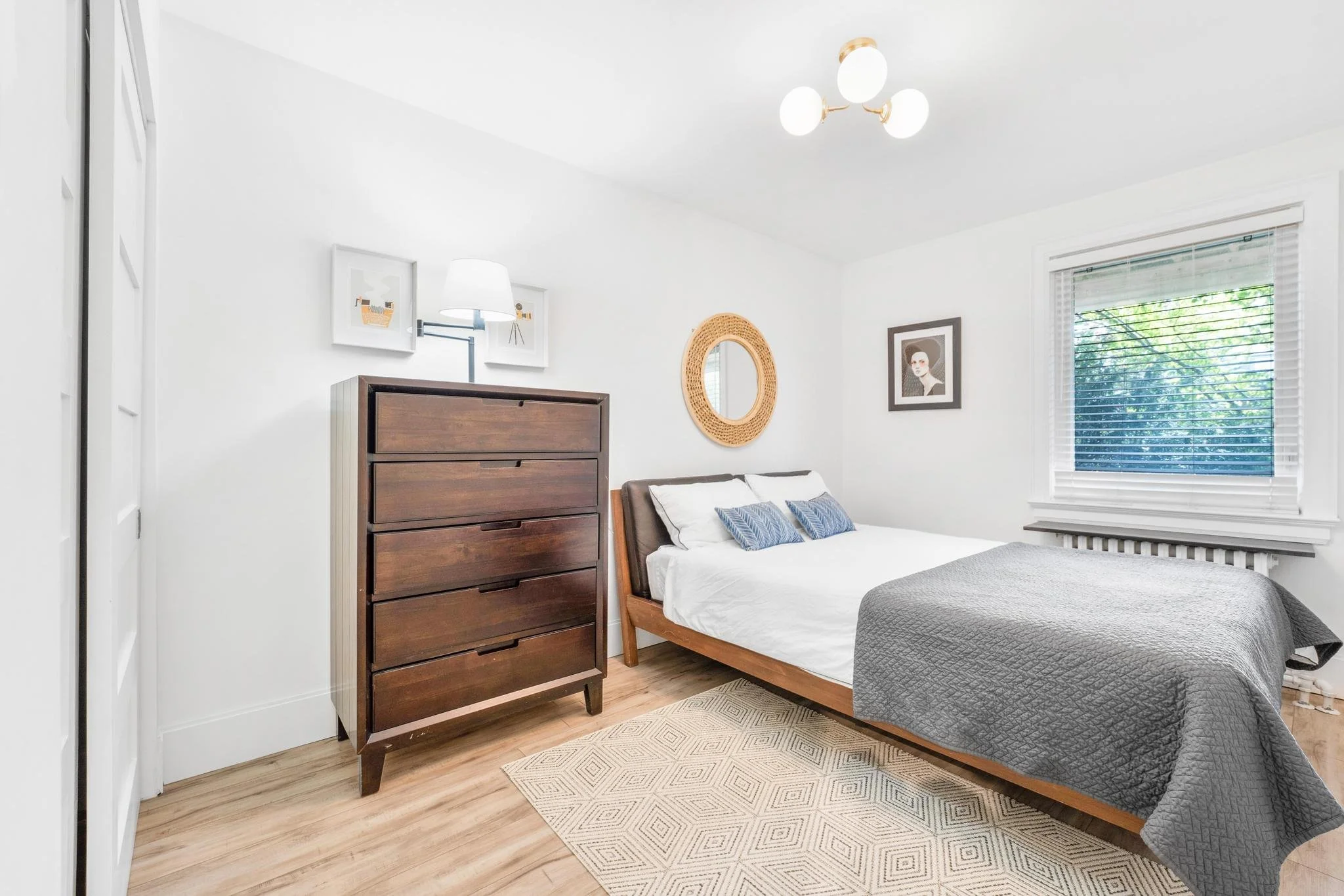 A bright bedroom with a wooden bed frame, white bedding, and gray quilt. There's a dark wooden dresser, a round wicker mirror, framed artwork on the wall, a window with blinds, and a modern ceiling light fixture.