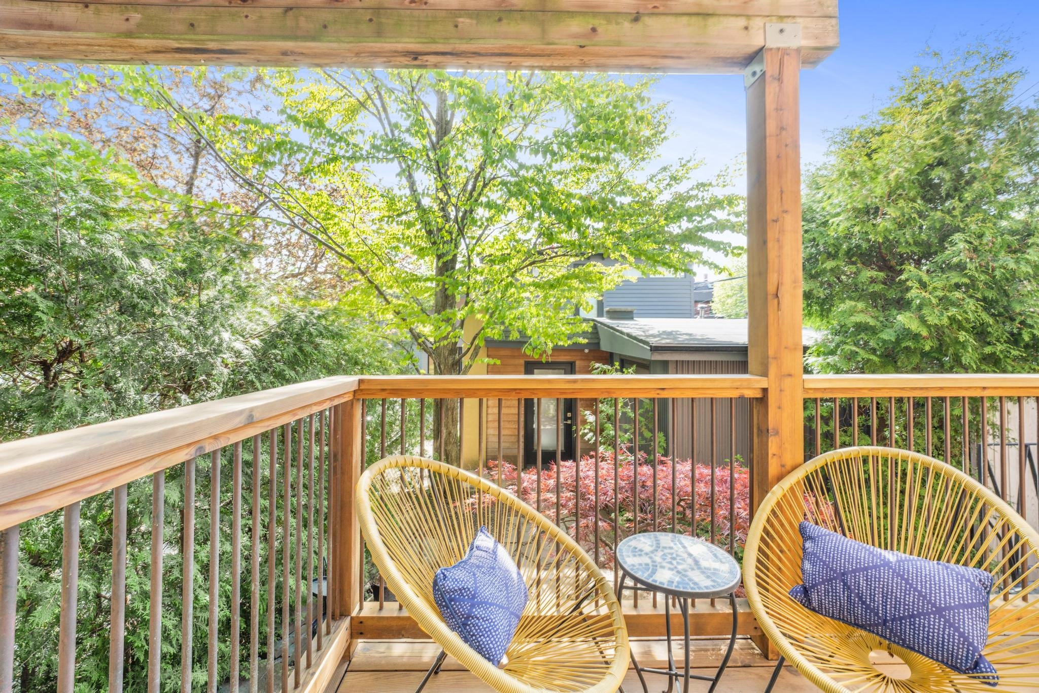 Balcony with two yellow Acapulco chairs with blue patterned cushions and a small round black table, overlooking lush green trees and a building with glass doors.