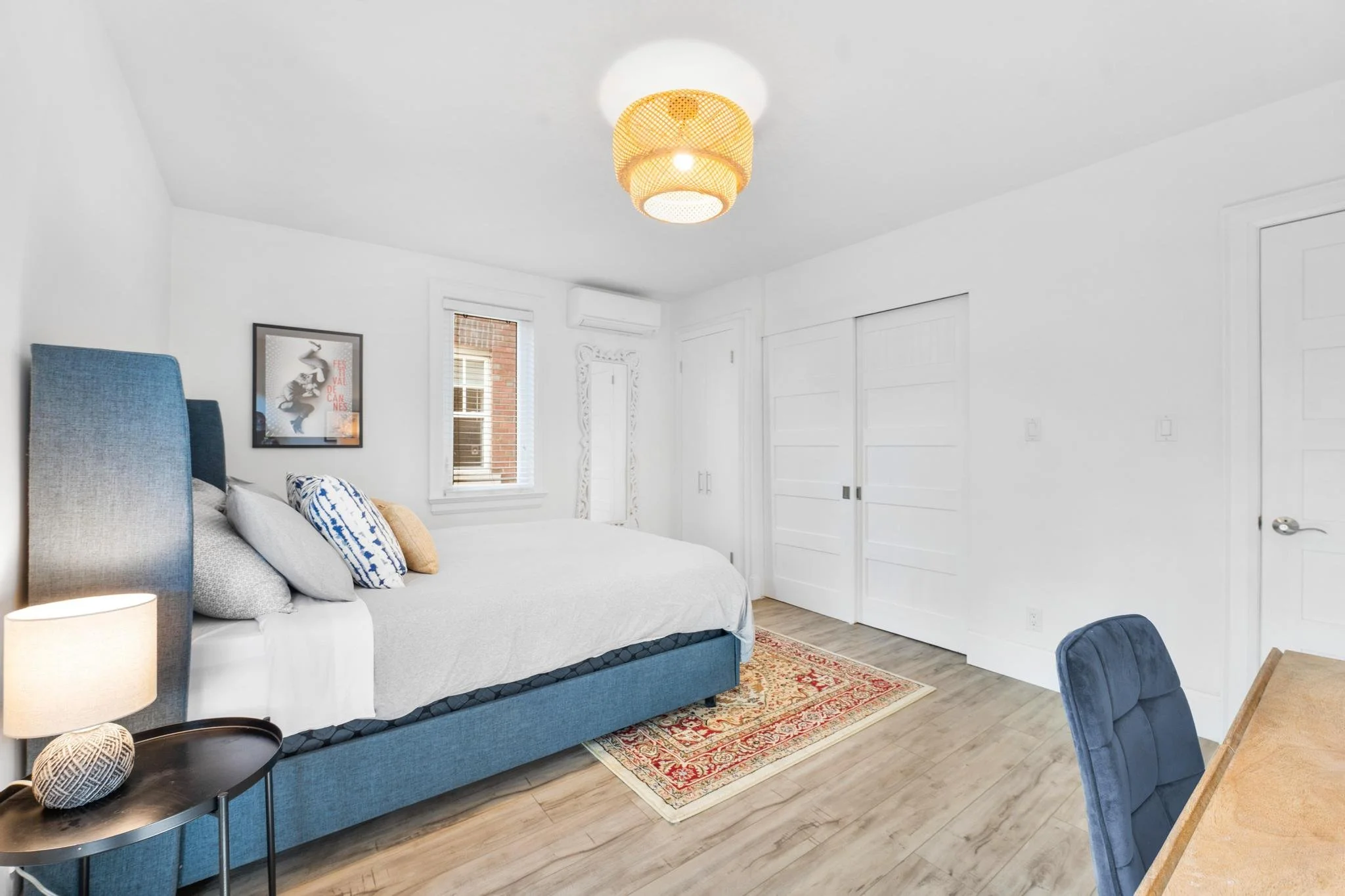 Bright bedroom with white walls, a blue bed with pillows, a small side table with a lamp and decorative object, a window, white closet doors, a colorful rug, and a blue upholstered chair near a wooden table.