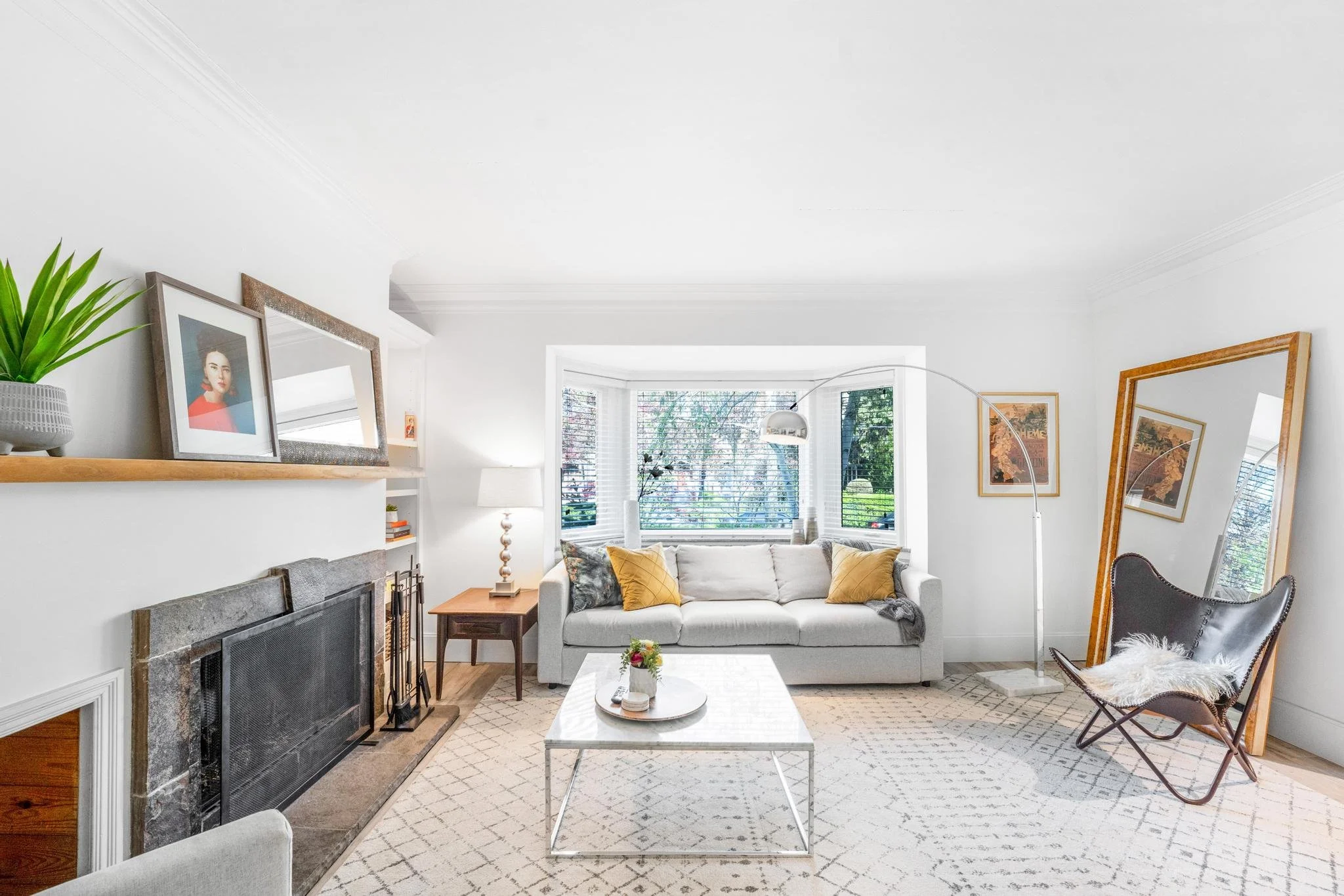 Bright living room with white walls, gray sofa with yellow pillows, a large mirror leaning against the wall, a marble coffee table, and a sitting chair with a sheepskin throw.