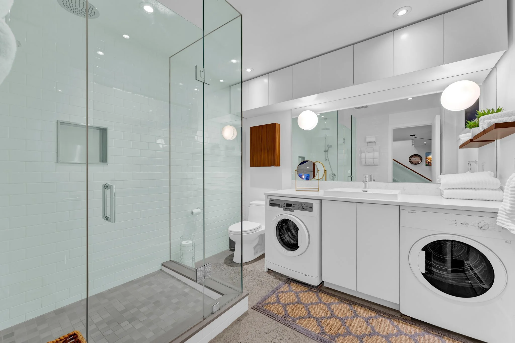 Modern bathroom with a glass shower on the left, white tile walls, a toilet, a washer and dryer under a white countertop, a large mirror, and a small decorative mirror on the counter. There are white shelves with potted plants and towels, and the roo