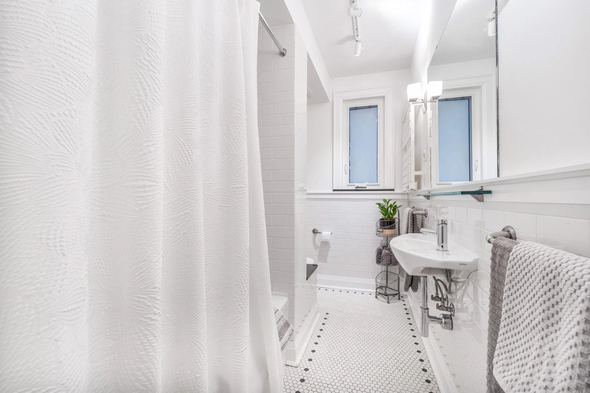 Bright white bathroom with a small pedestal sink, wall-mounted towel racks with gray towels, a window, and a shower with a textured white curtain.