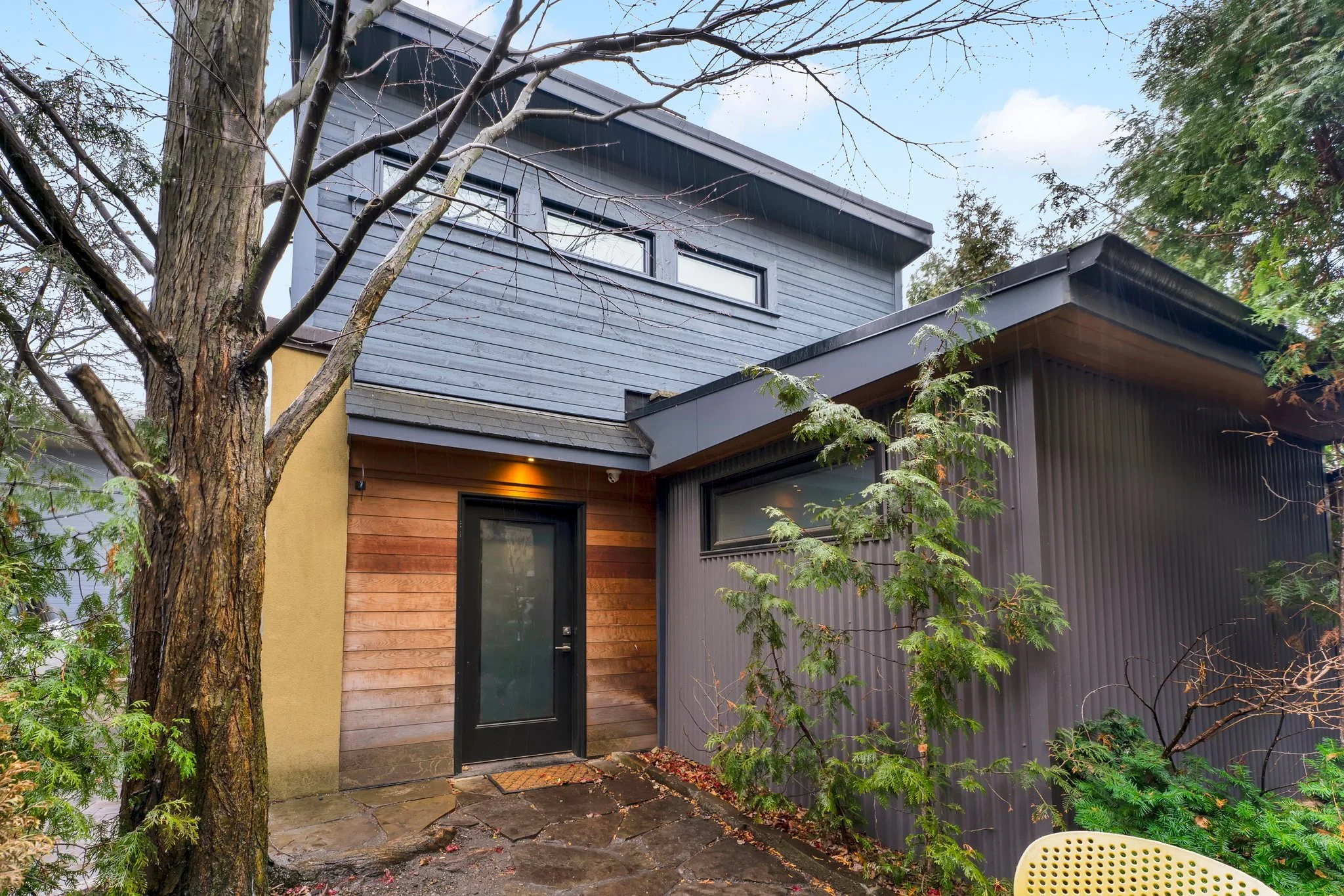 Exterior of a modern house with gray wooden and black metal siding, a black door, and a small stepped stone pathway, surrounded by trees and plants.