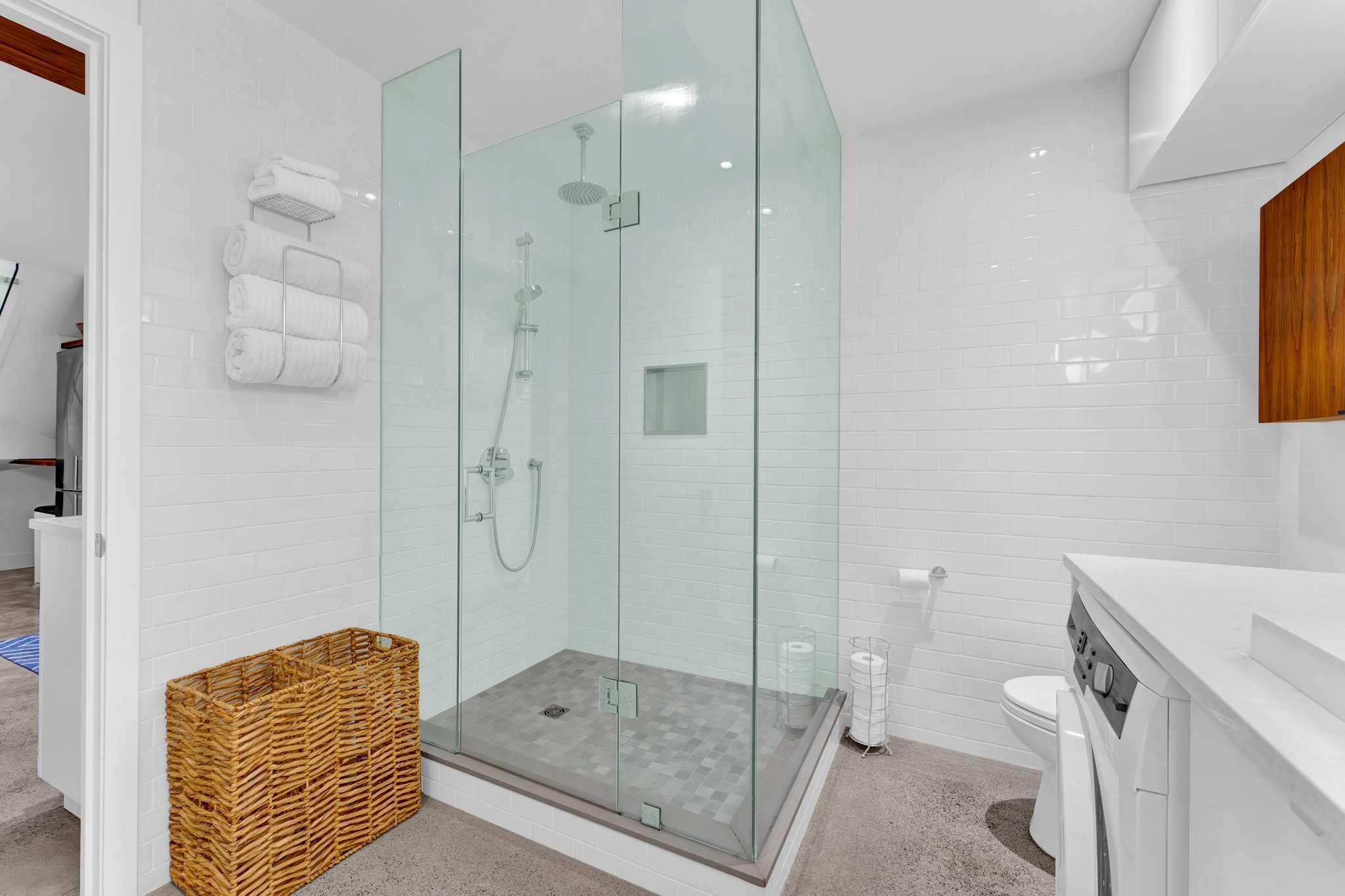 Modern bathroom with a glass shower enclosure, white tiled walls, a wooden cabinet, a washing machine, and a wicker laundry basket.