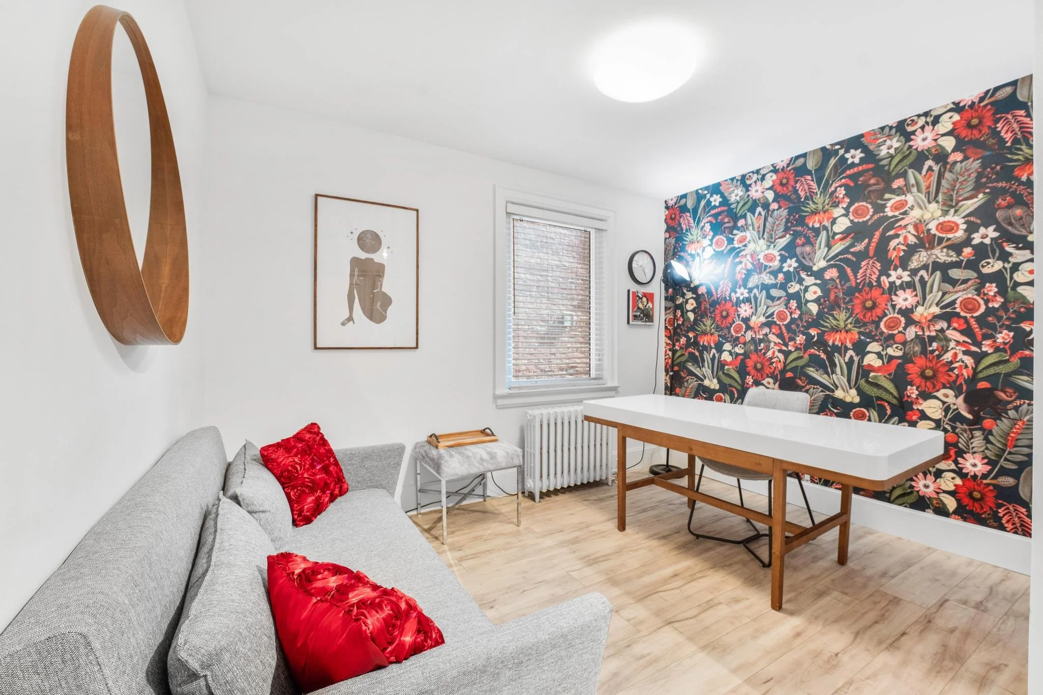 Living room with a gray sofa with red pillows, a table, a floral wallpapered accent wall, a window with blinds, and minimal decor.