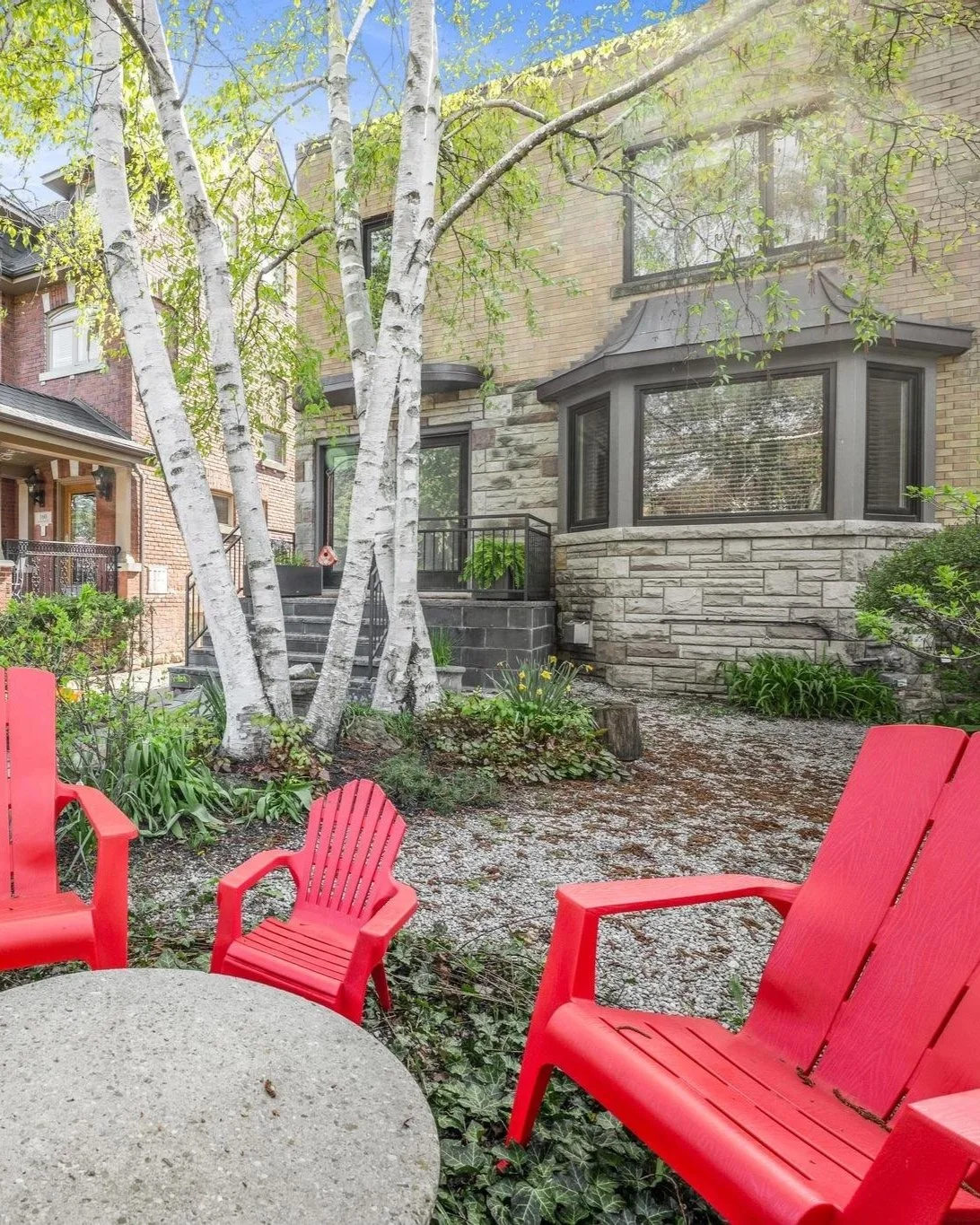 View of a landscaped backyard patio area with red Adirondack chairs and a round concrete table, surrounded by trees, plants, and a two-story brick house with bay windows and a small set of stairs leading to a deck.