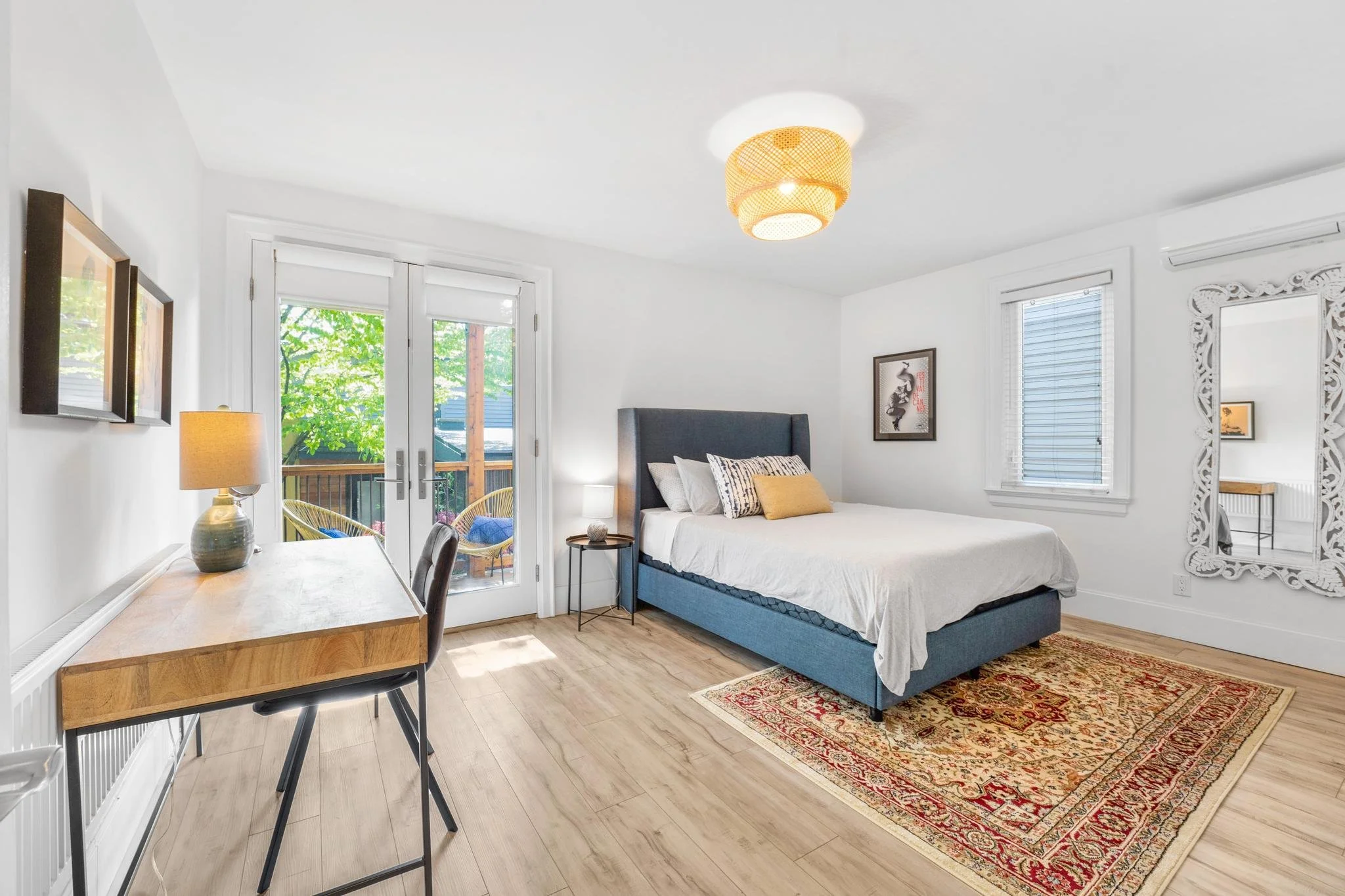 Bedroom with a blue upholstered bed, white bedding, a carved white mirror, a wooden desk with a lamp, and glass doors leading to a balcony with chairs and trees outside.