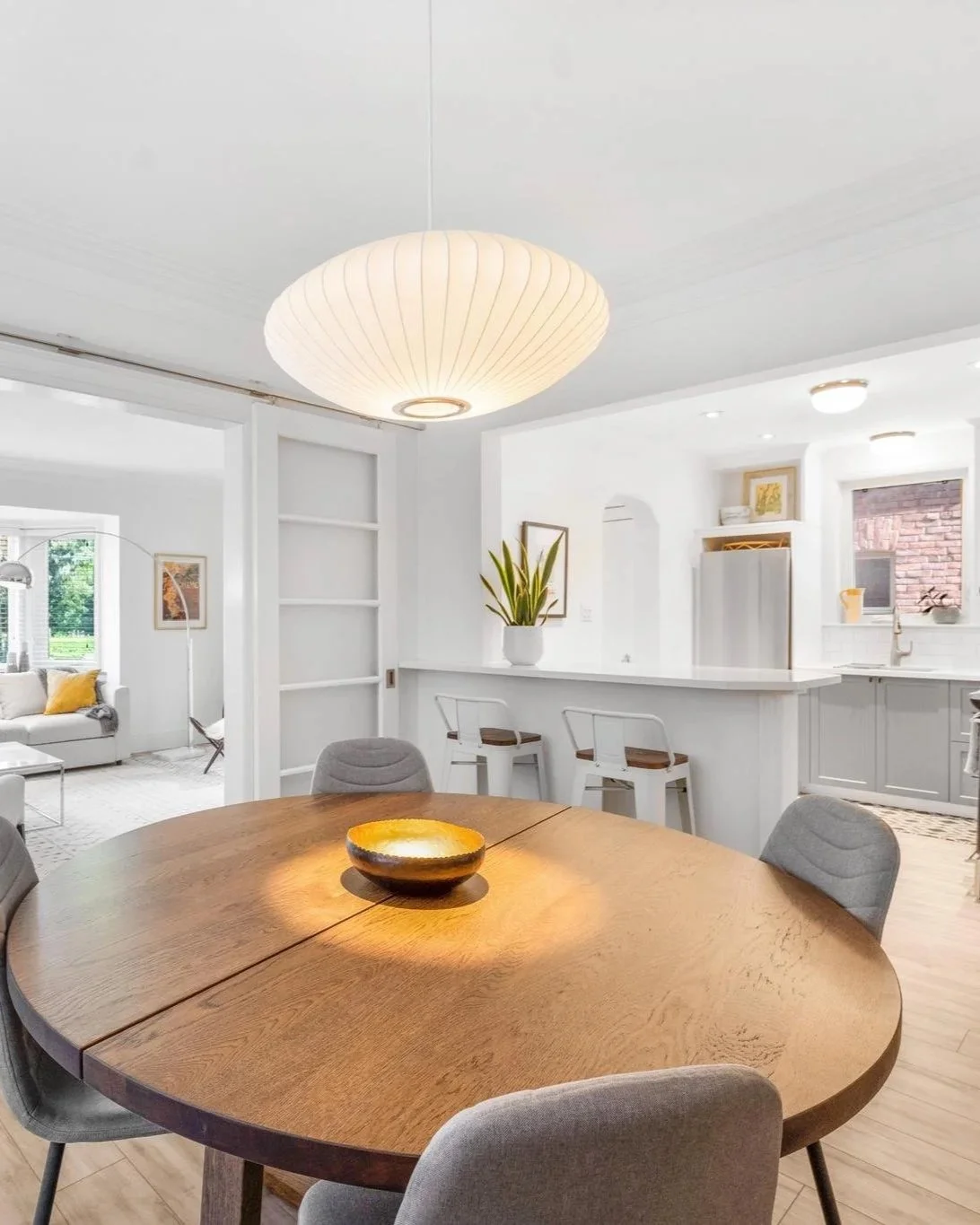 A modern dining room with a round wooden table, gray chairs, and a white pendant light hanging from the ceiling. The background shows a white kitchen with bar stools, a potted plant, and bright windows.