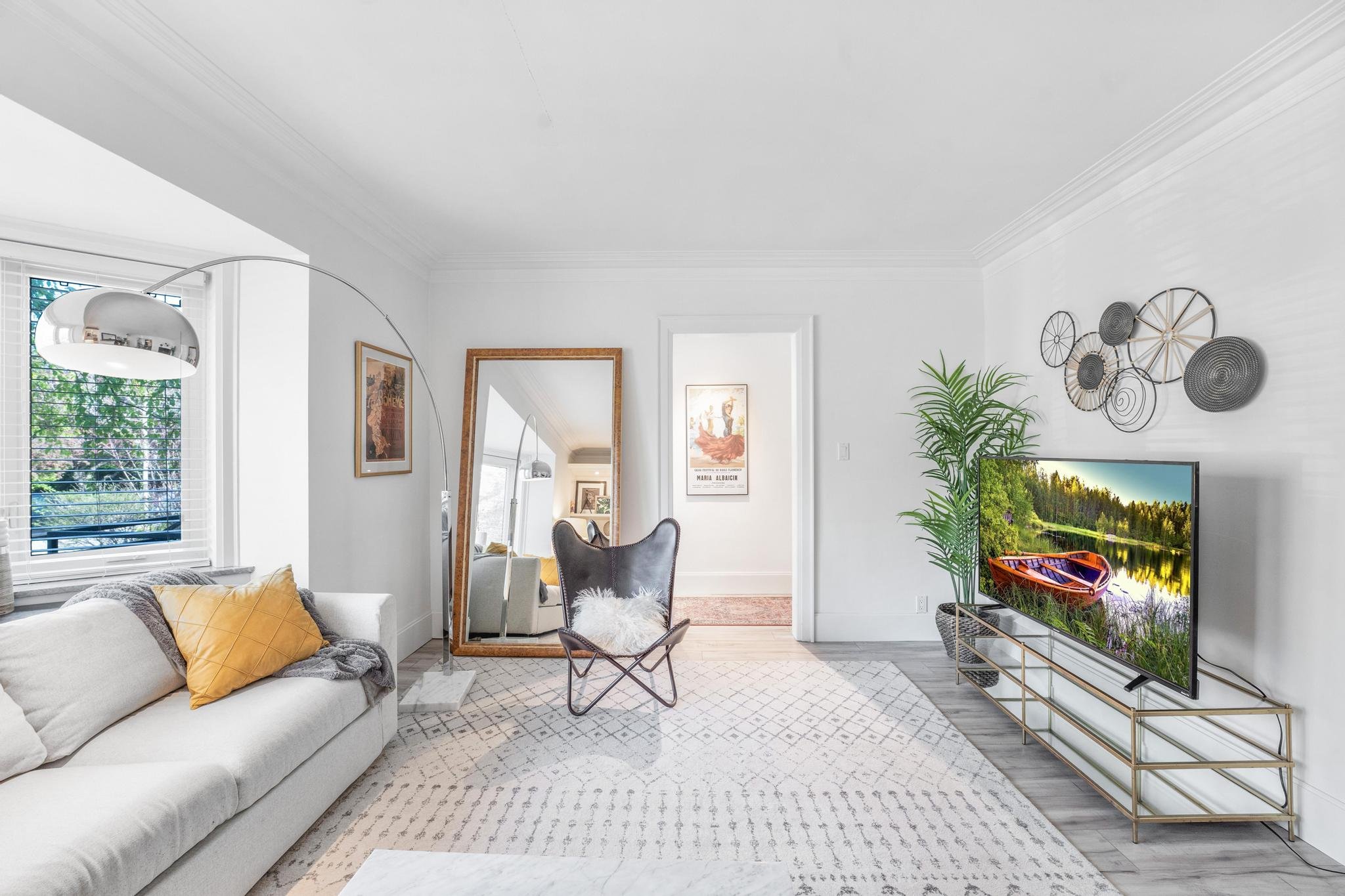 Bright living room with a large window, white sofa with yellow and gray pillows, a full-length mirror, black chair with a white fluffy pillow, a TV on a gold metal stand, a potted plant, and wall decor.