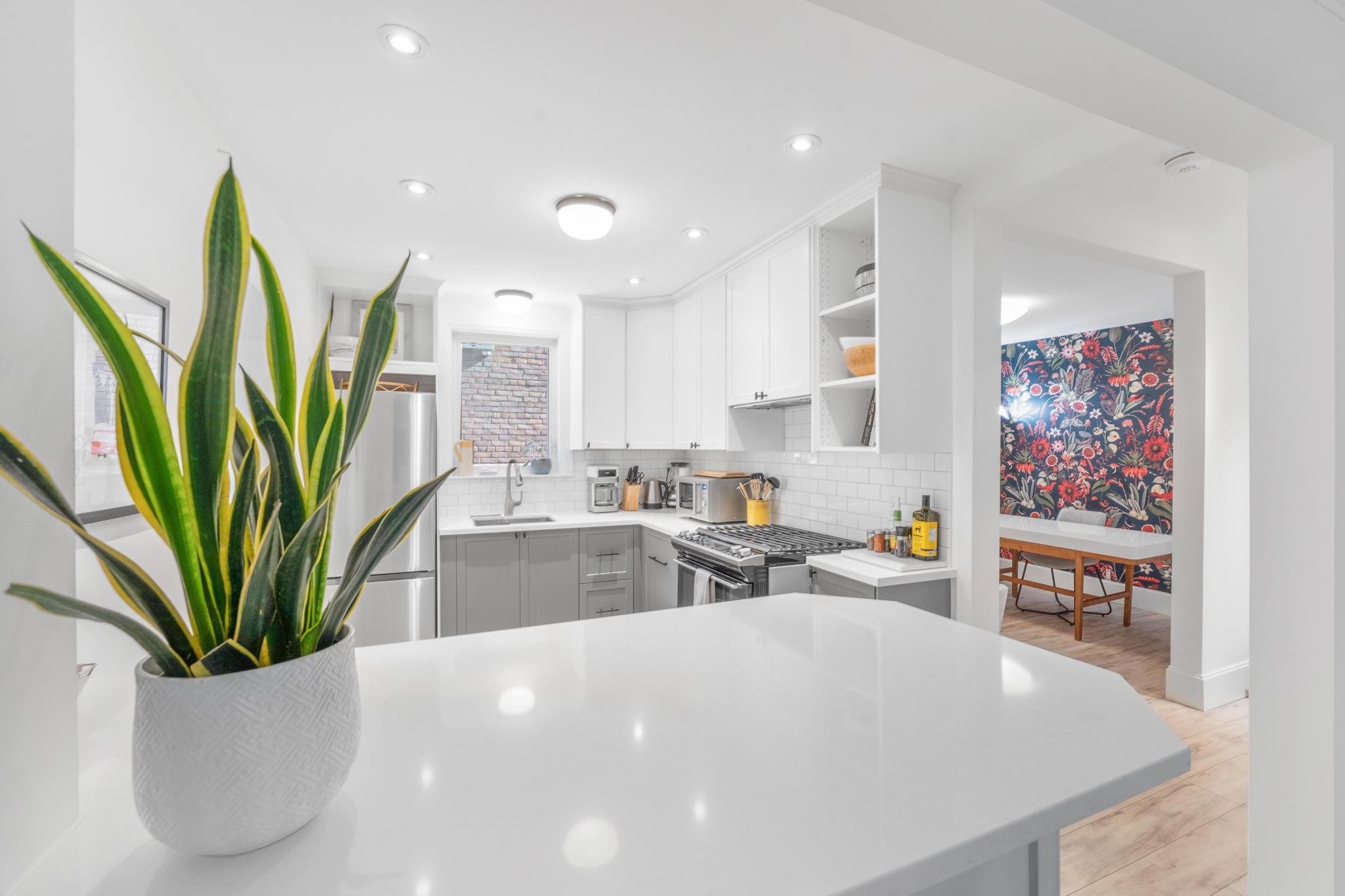 Modern white kitchen with gray lower cabinets, white upper cabinets, stainless steel appliances, a white subway tile backsplash, and a window over the sink, with a plant in the foreground and a floral wallpaper accent wall in the dining area.