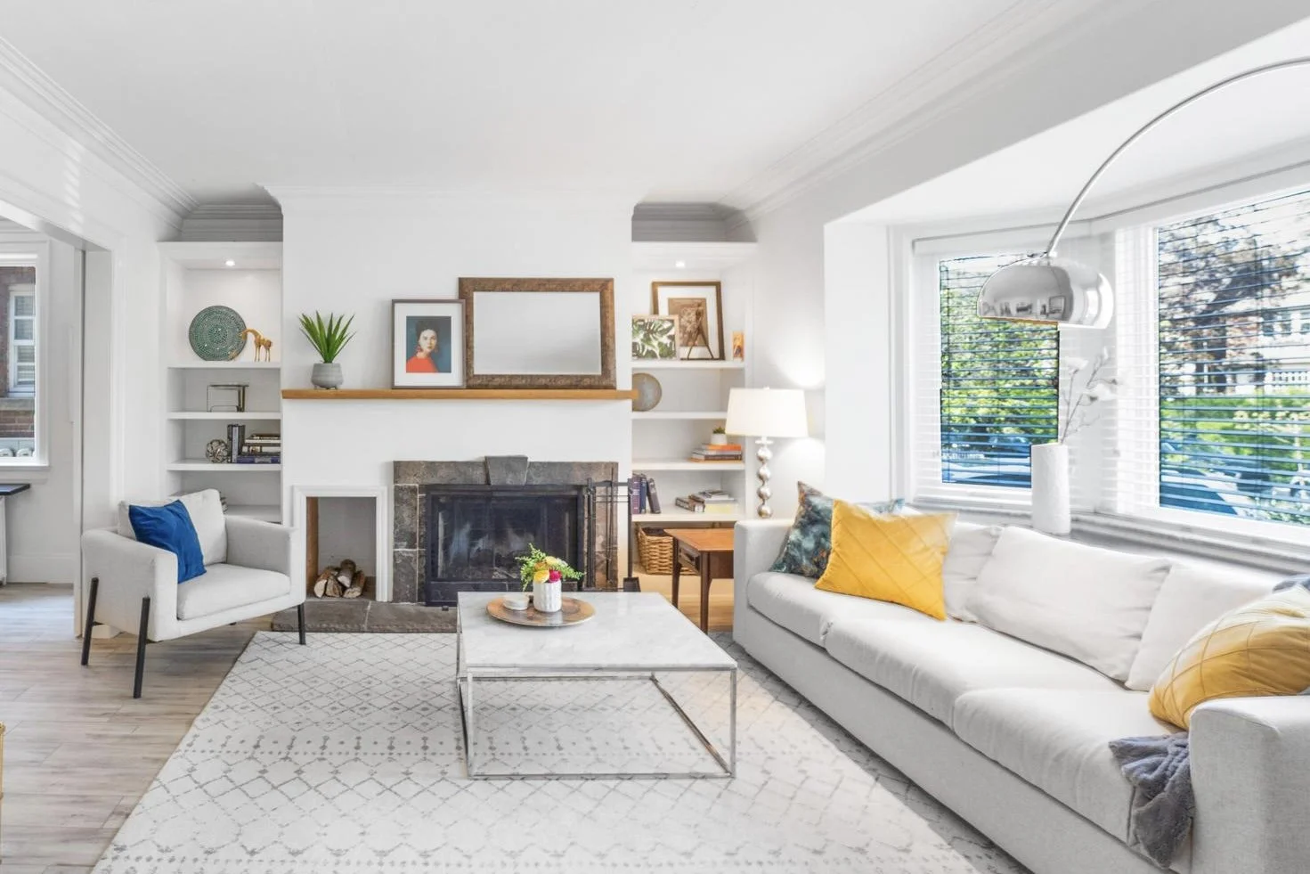 Living room with white sofa, yellow pillows, and a glass coffee table, fireplace with mantle, built-in white bookshelves, large window with blinds, and modern floor lamp. Decor includes framed art, books, and plants.