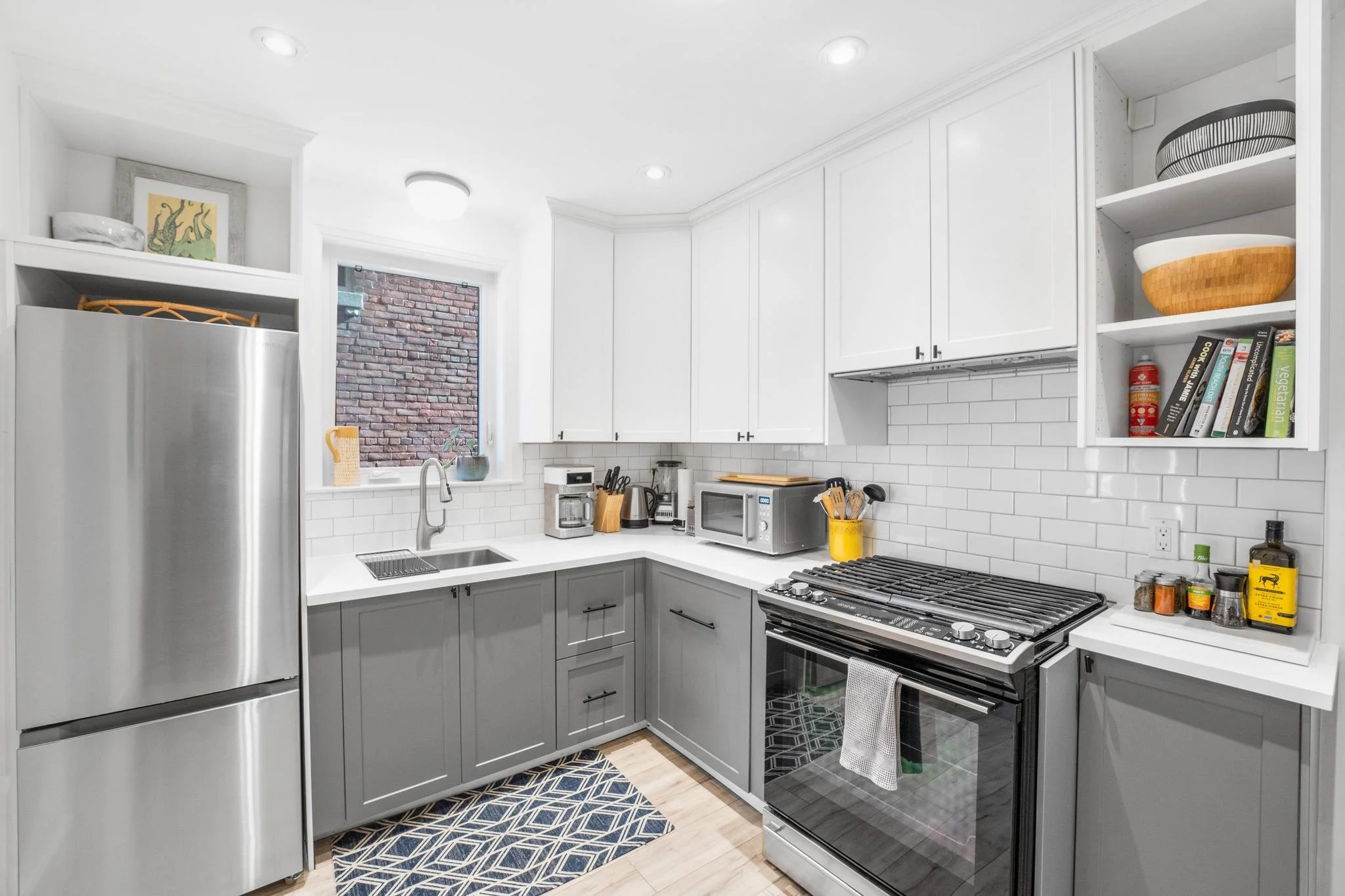 Kitchen with white and gray cabinets, stainless steel refrigerator, microwave, coffee maker, toaster, spice jars on a white countertop, and open shelves with bowls, cookbooks, and kitchen essentials.