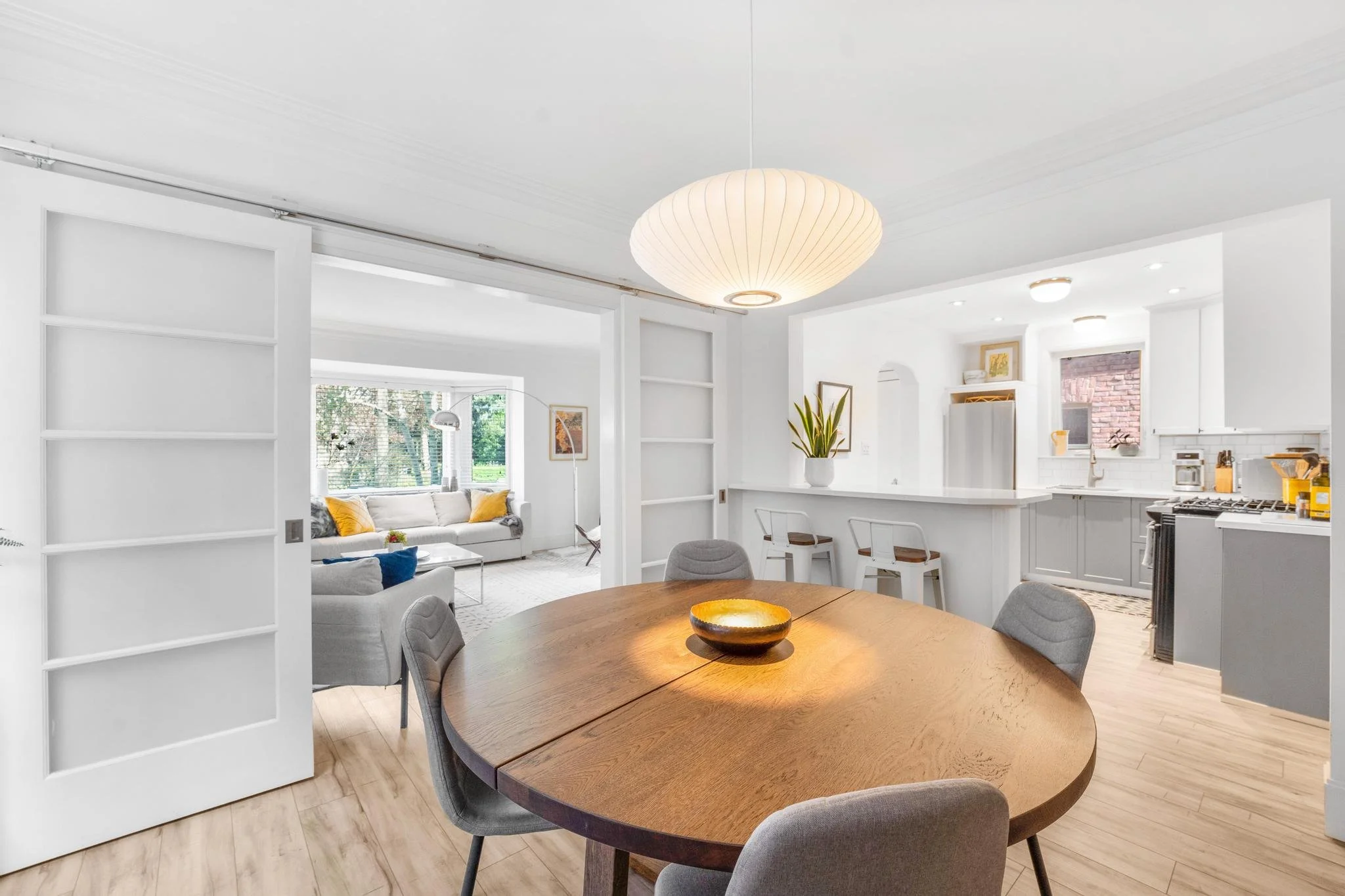 A bright, open-concept dining and living room with white walls, light wood flooring, a wooden dining table with grey chairs, and a white kitchen with grey cabinets. There is a large window in the living room with a white sofa and yellow pillows, and 