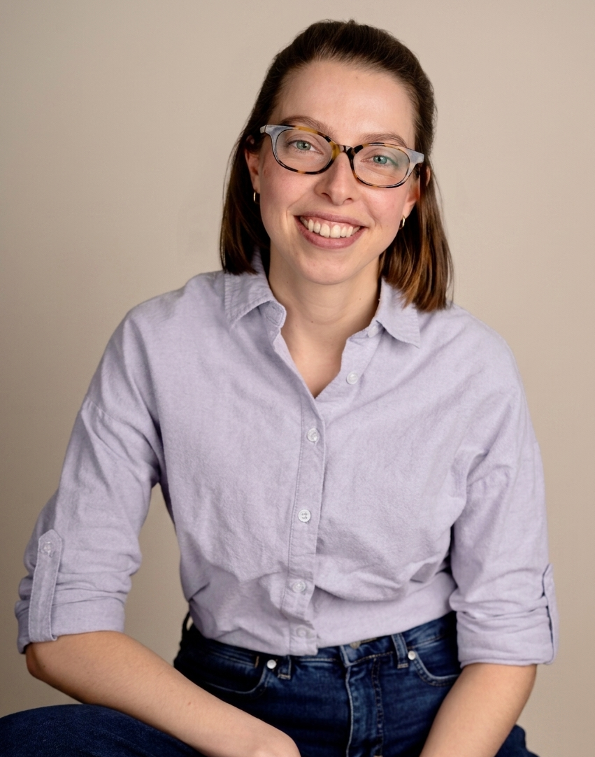 Photo of Kim Vassallo, Psychologist (MSc) and Gestalt therapist (in training) with shoulder-length brown hair, wearing glasses and a light blue button-up shirt, smiling at the camera against a plain background.