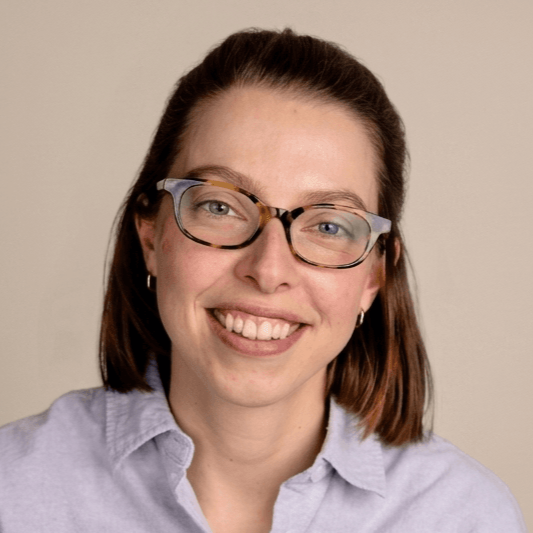 Profile picture of Kim Vassallo, psychologist and gestalt therapist in training. She has shoulder-length brown hair, glasses, and a light blue button-up shirt smiling at the camera against a plain beige background.