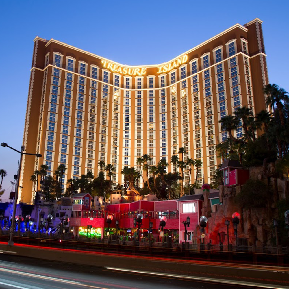 Nighttime view of the Treasure Island hotel and casino in Las Vegas with illuminated sign, tall building, palm trees, and colorful lights.