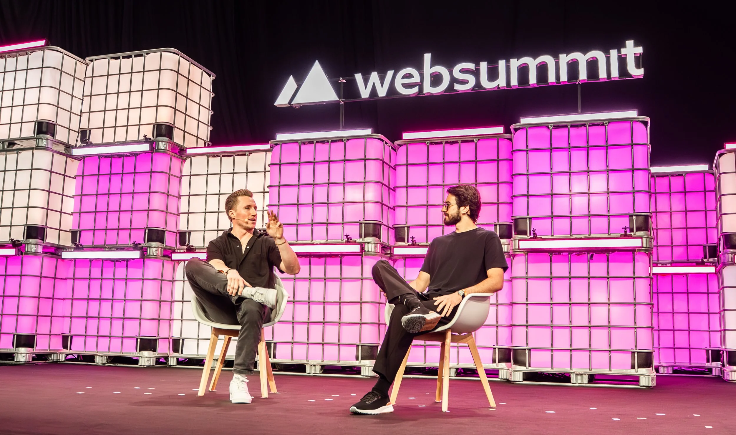 Two men seated on chairs facing each other on a stage with a large illuminated backdrop of pink and white cubes and the 'websummit' logo above.