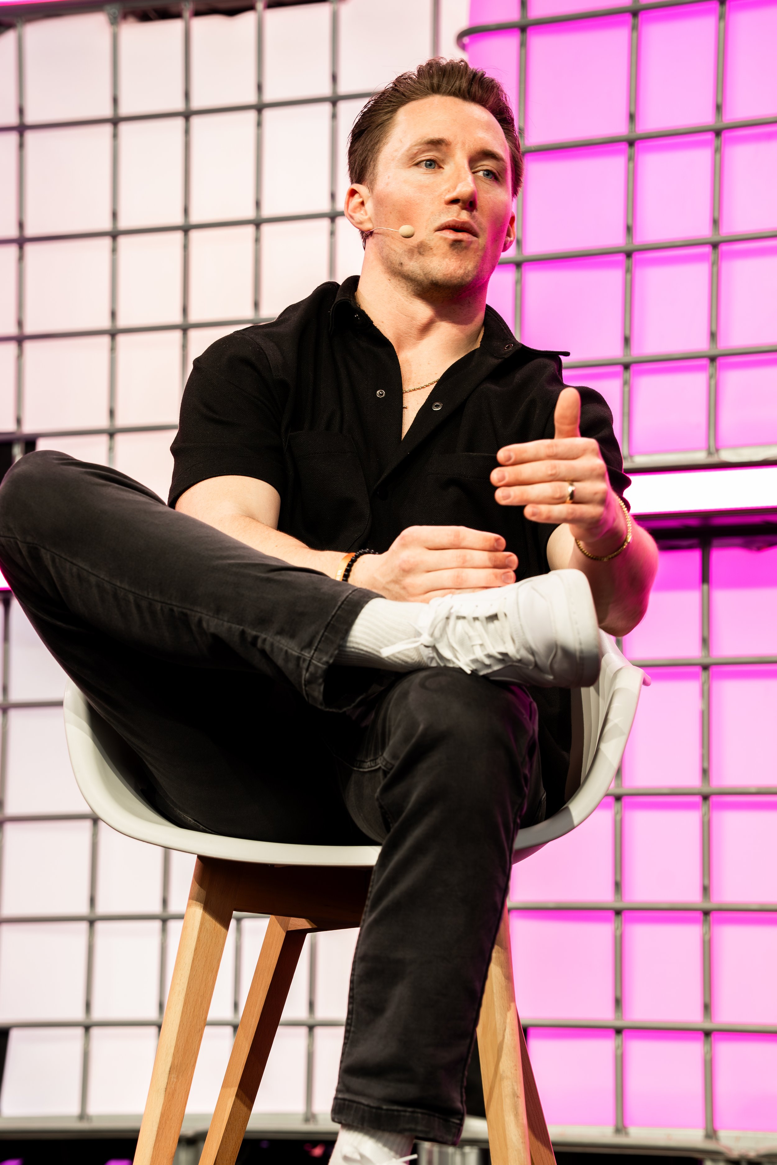 A man sitting on a white chair with crossed legs, wearing a black shirt, black pants, white shoes, and white socks. He has a microphone headset and is gesturing with his hands during a presentation or discussion against a background of pink and white panels.