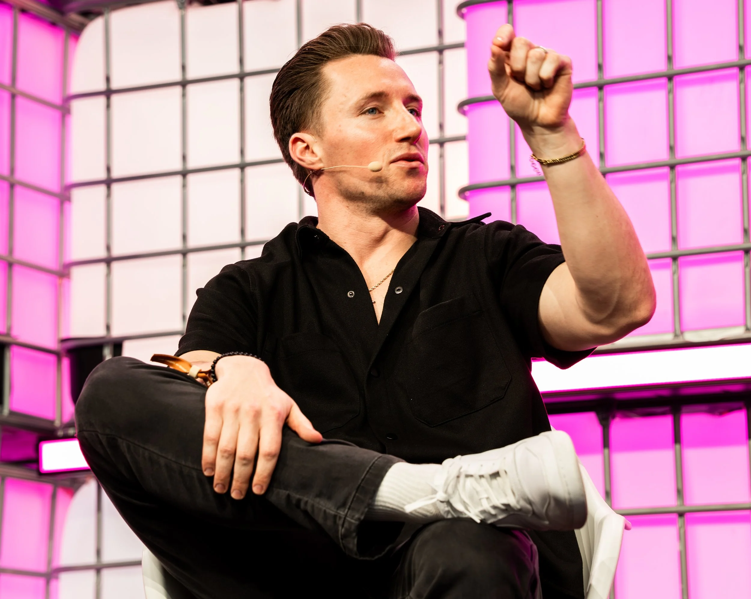 A man with styled hair and a microphone headset sitting cross-legged on stage, with a backdrop of pink and black grid panels, gesturing with his right hand.