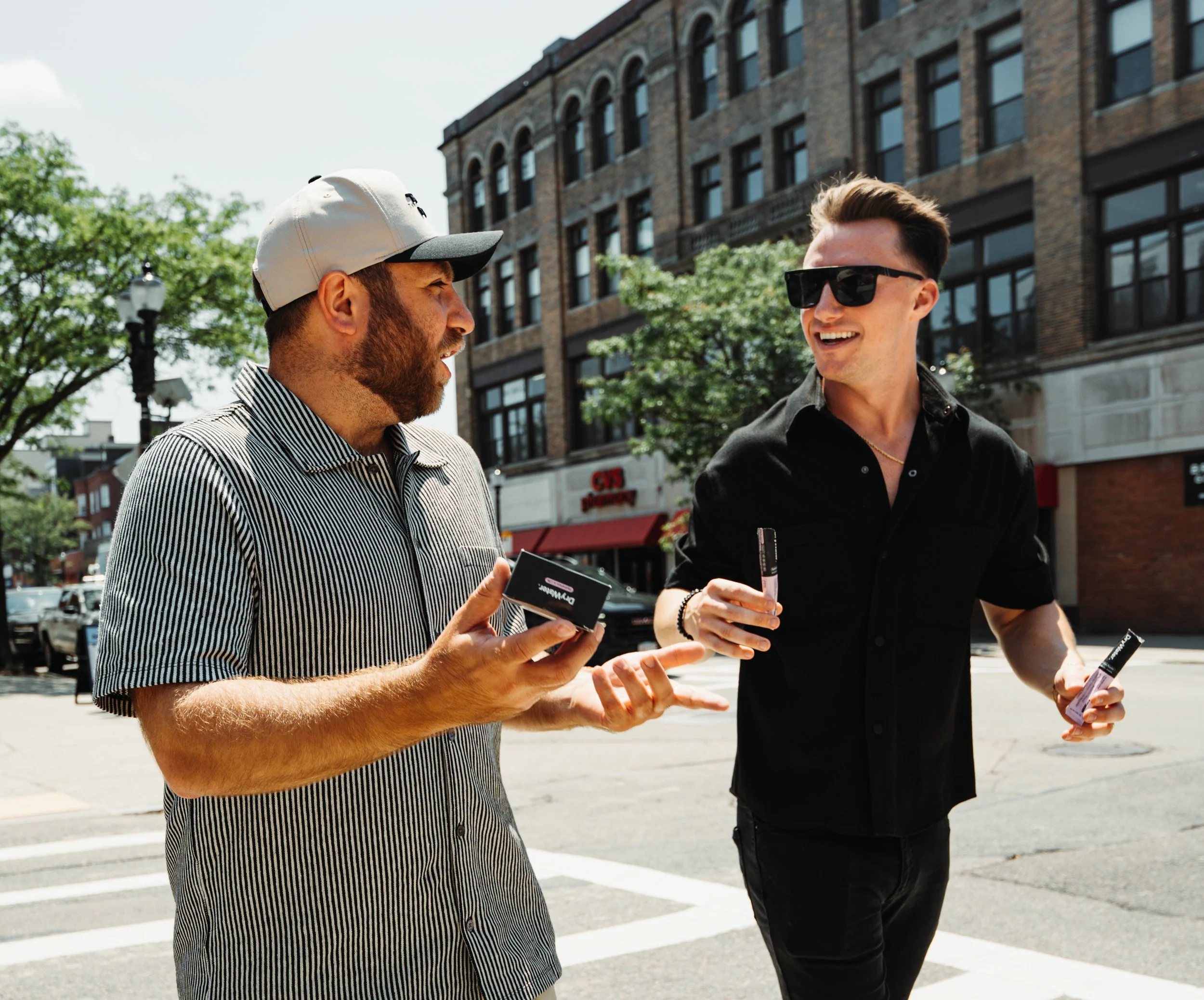Two men walking on a city street, talking and smiling, one wearing sunglasses and black shirt, the other with a cap and striped shirt, holding Lipton tea bottles.