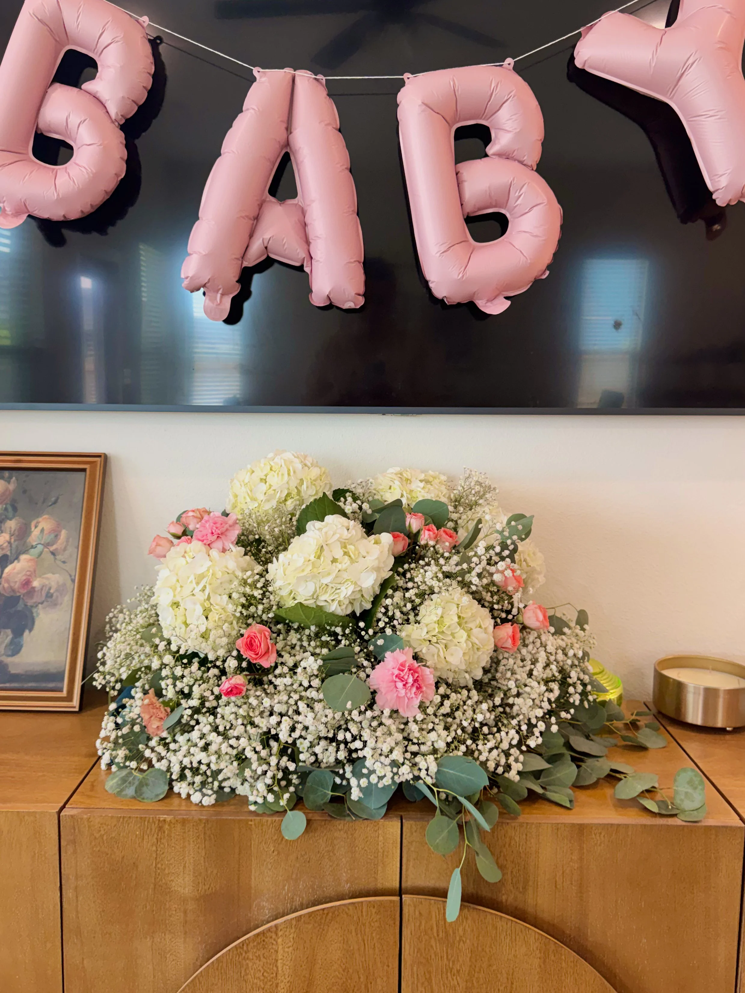 Pink inflated letter balloons spelling 'BABY' hanging above a large bouquet of white and pink flowers on a wooden surface.