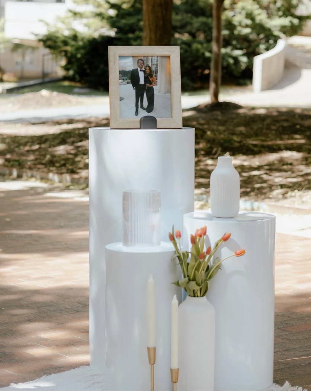 A engagement proposal display with a framed photo of a couple dressed formally, set on a white pedestal with vases, candles, and flowers in an outdoor setting with trees and sunlight.