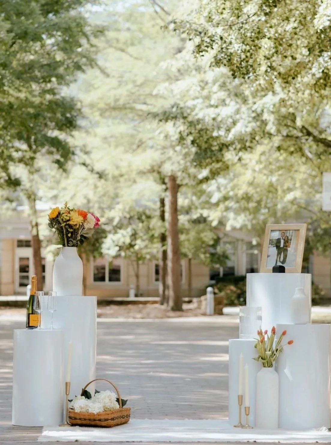 Outdoor wedding or event setup with white cylindrical pedestals, vases with flowers, a framed picture, and a basket of flowers, with green trees in the background.