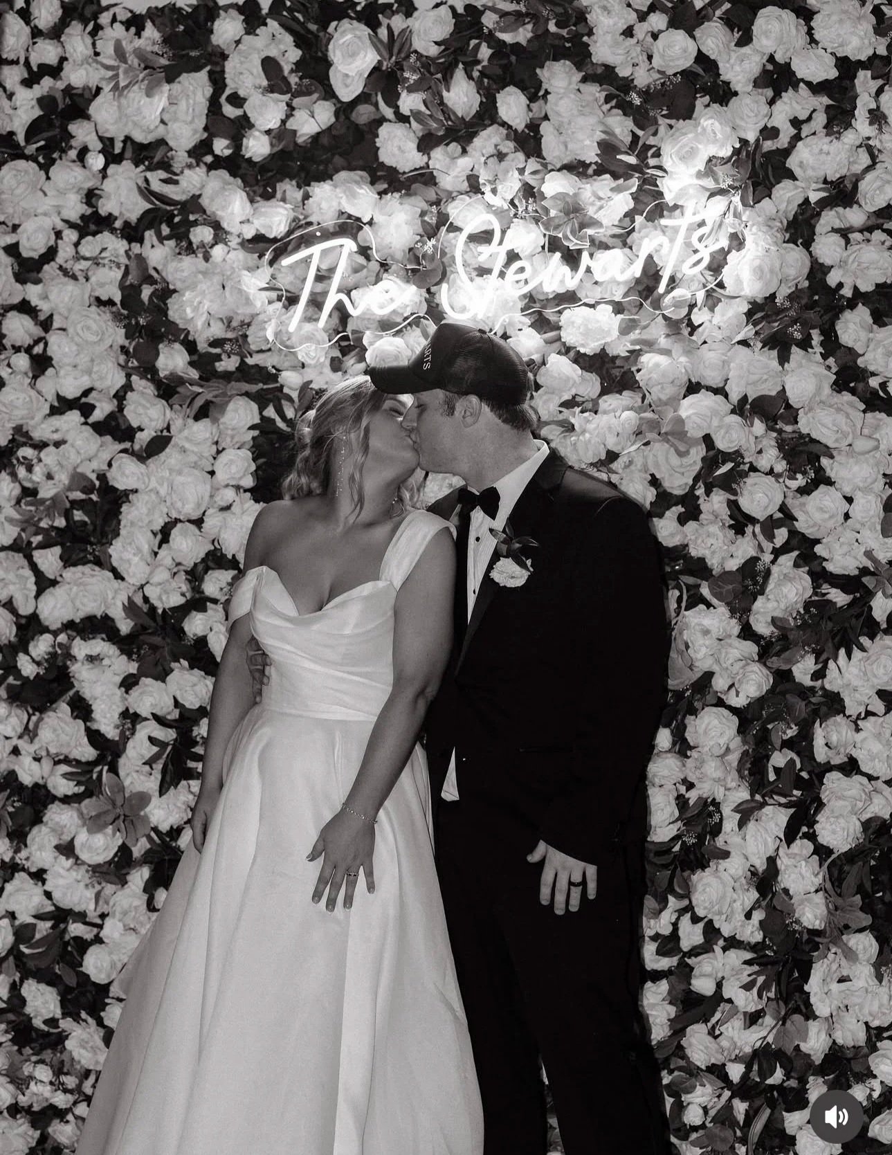 A couple in wedding attire kissing in front of a floral backdrop with a neon sign that reads 'The Chewats'. The bride wears a strapless white gown, and the groom wears a tuxedo with a bow tie and a cap.