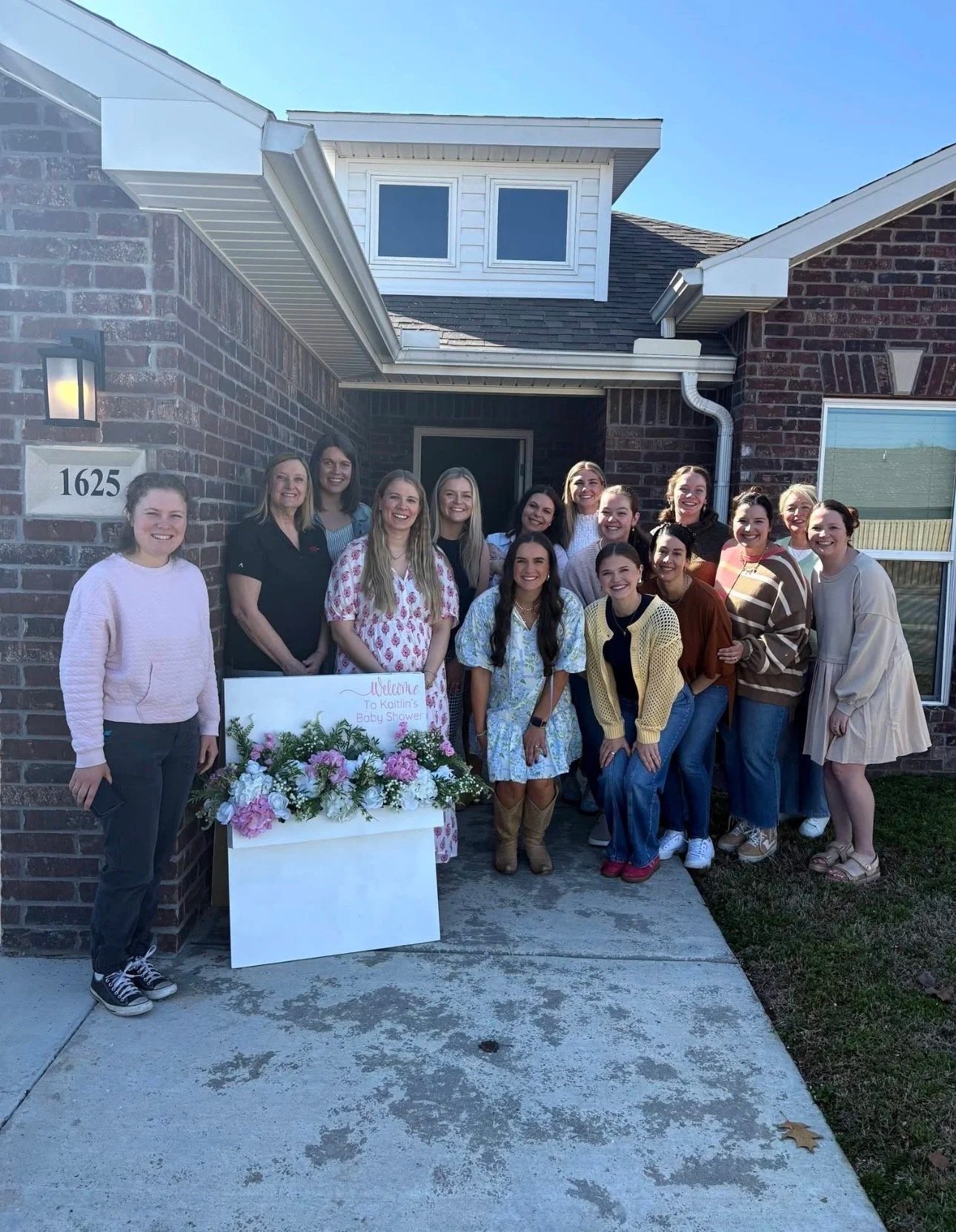 Group of women standing outside a brick house for a baby shower, with a floral display and a welcoming sign.