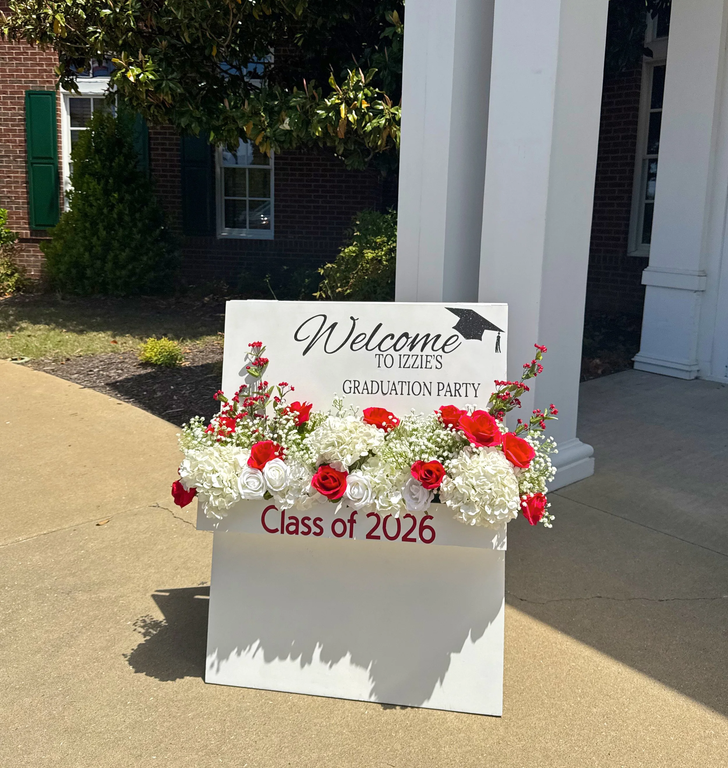 A floral display in front of a white sign that reads "Welcome to Izzi's Graduation Party Class of 2026" with red and white flowers arranged at the base, placed outdoors on a sidewalk.